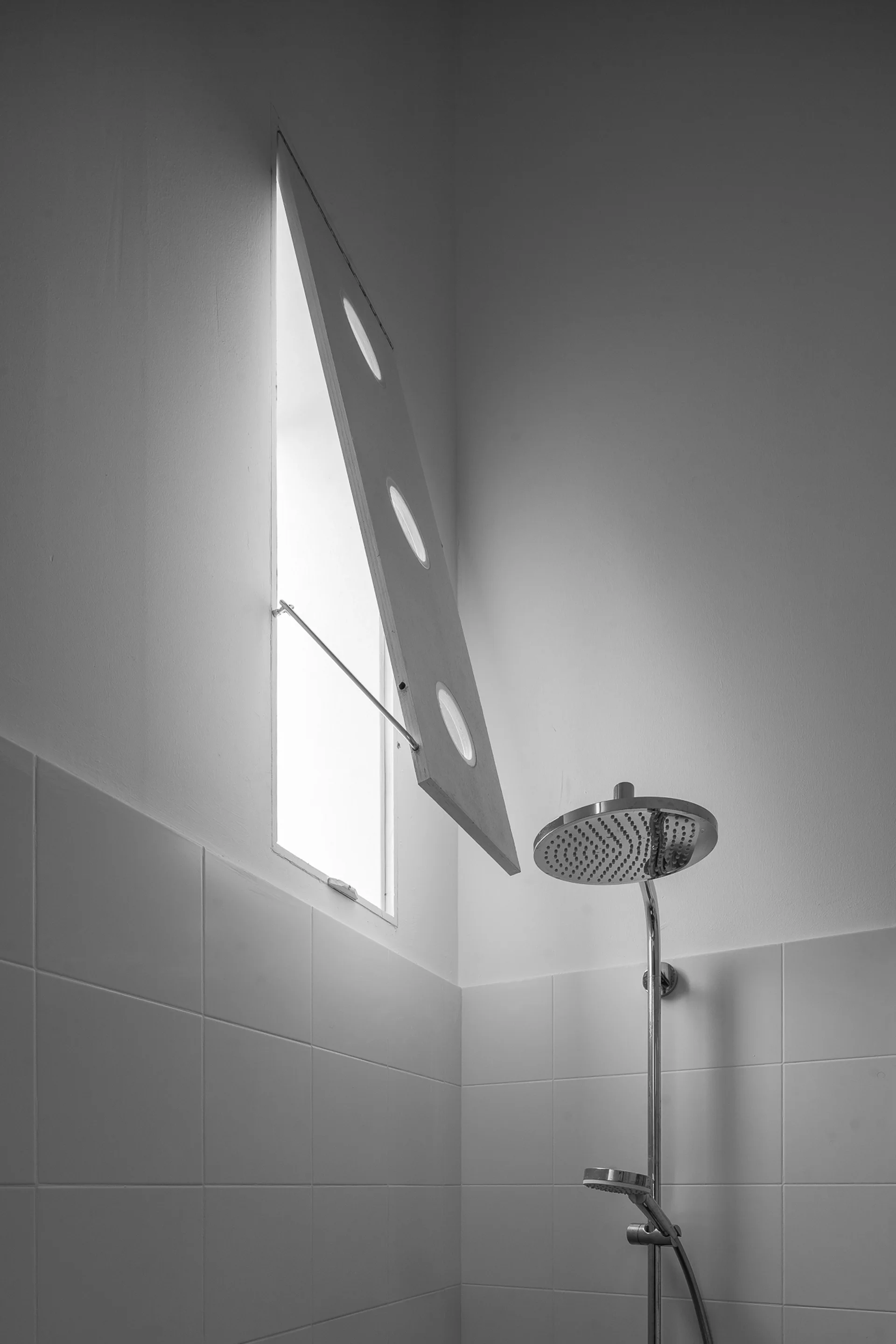 A white tiled bathroom shower corner features a chrome rainfall shower head and a triangular hinged window with three circular glass cutouts.