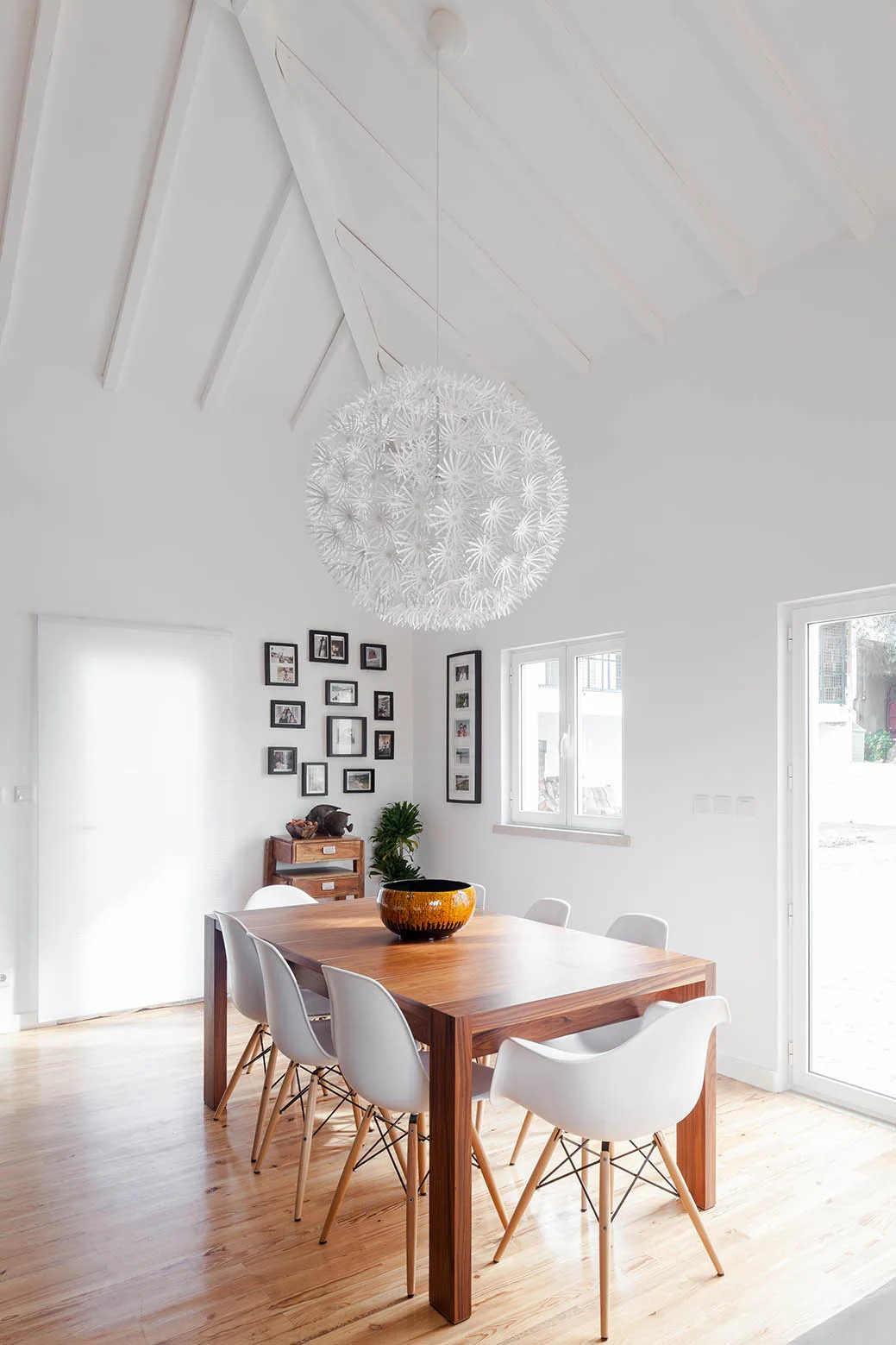 A wood dining table is surrounded by white chairs under a large spiky white pendant light and a white vaulted ceiling.
