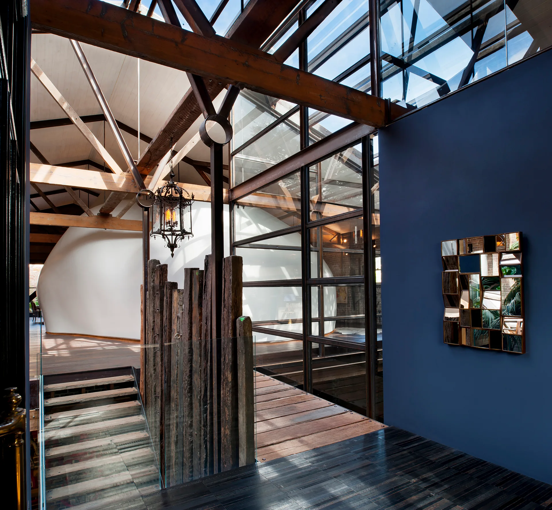 A modern home landing features exposed wood ceiling beams, a blue accent wall with a geometric mirror, and a staircase with vertical timber posts.