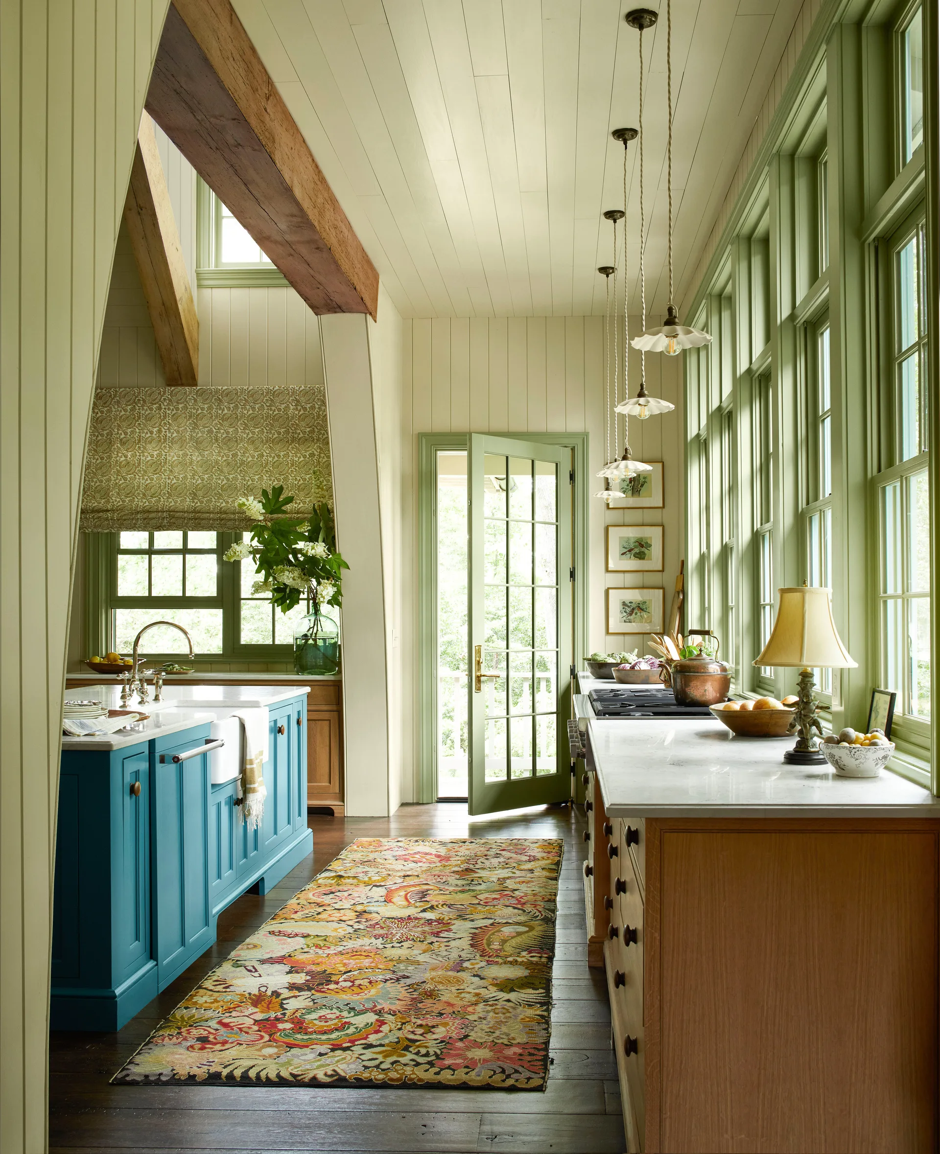 A bright cottage kitchen features a blue painted island with an apron sink, wood lower cabinets, and a long wall of sage green window frames.
