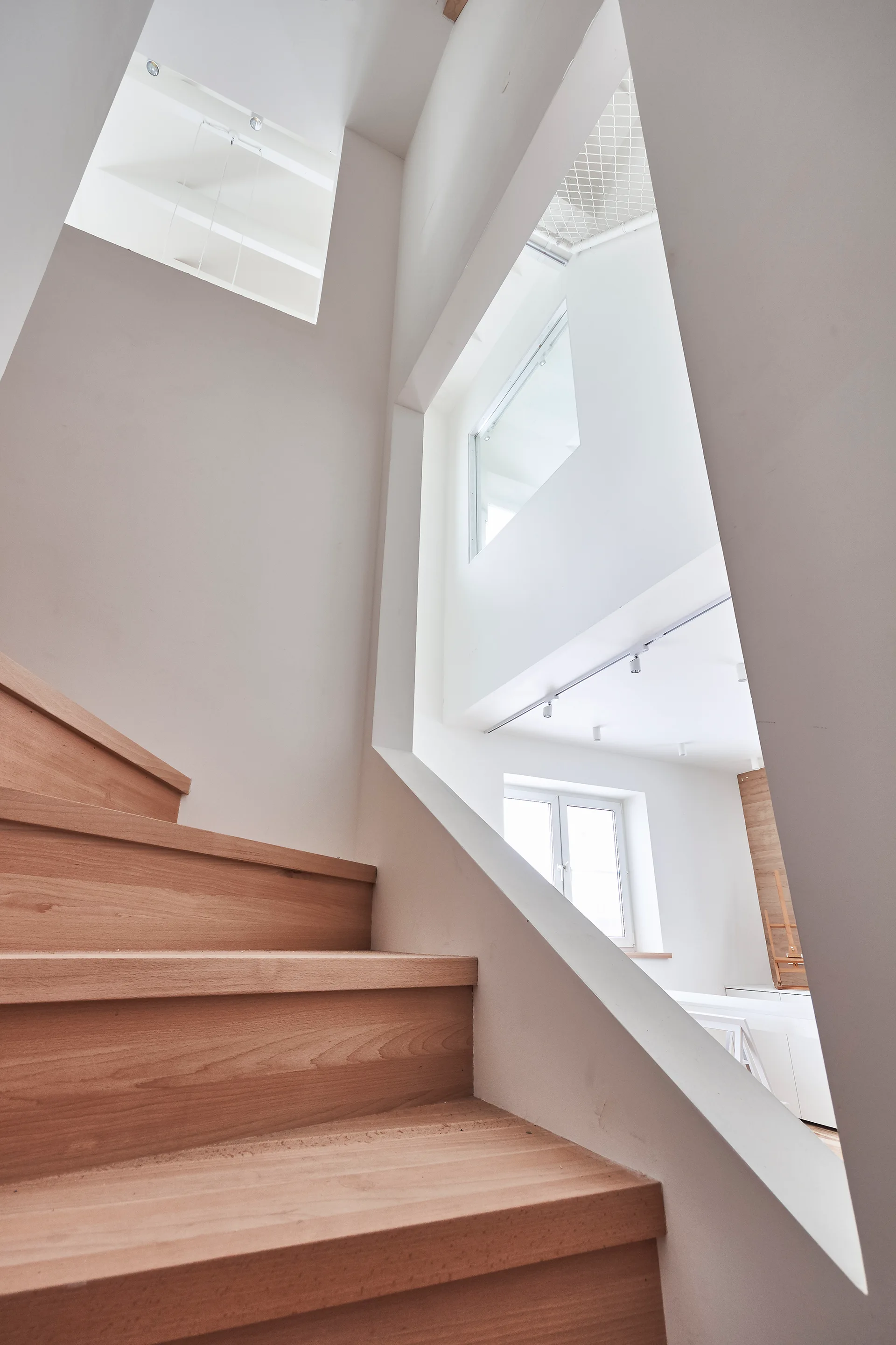 A modern wooden staircase with light brown steps rises against white walls featuring a large angular wall cutout.