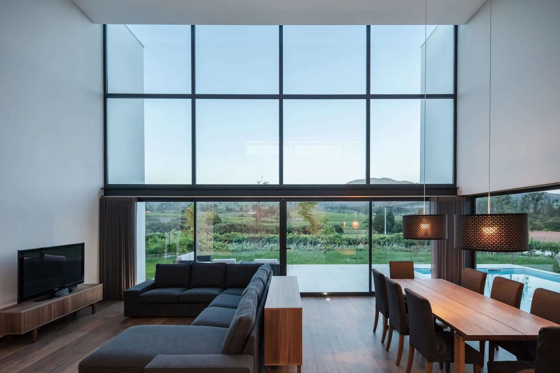A modern open-plan living and dining room featuring a grey sectional sofa, wooden dining table, and a massive wall of floor-to-ceiling windows.