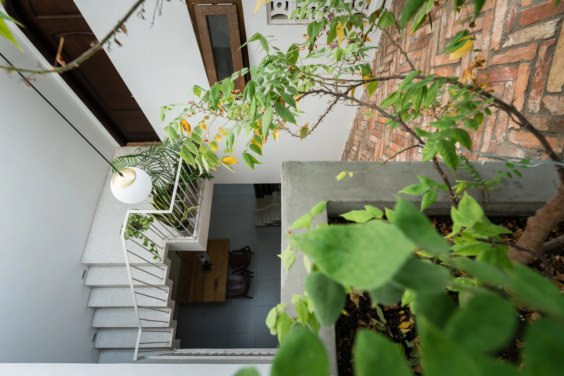 High-angle view looking down a white concrete staircase with a metal railing past a hanging spherical light and concrete planters toward a dining area.