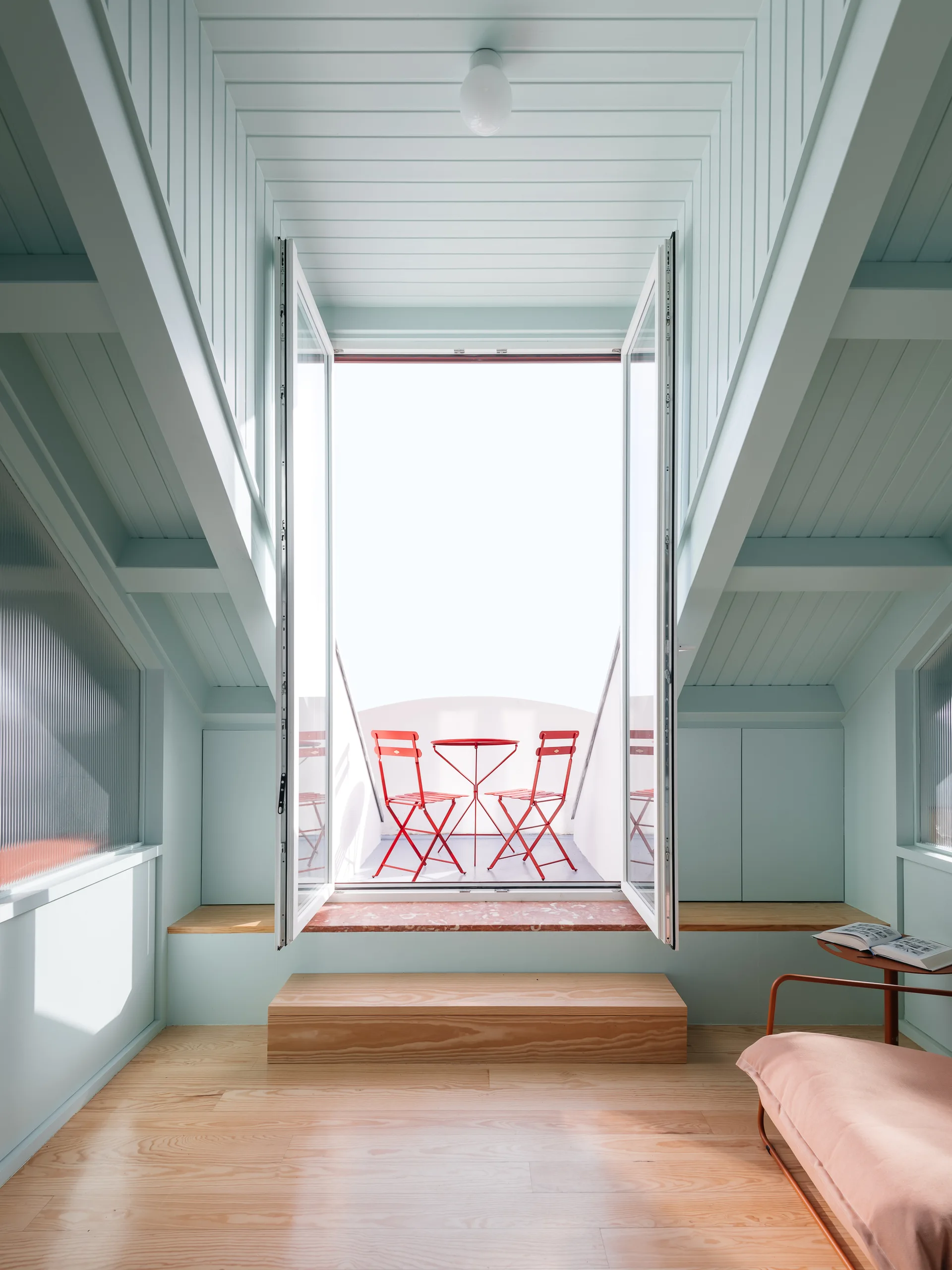 Open white French doors lead from a mint green attic room with wood floors to a balcony featuring a red metal bistro set.
