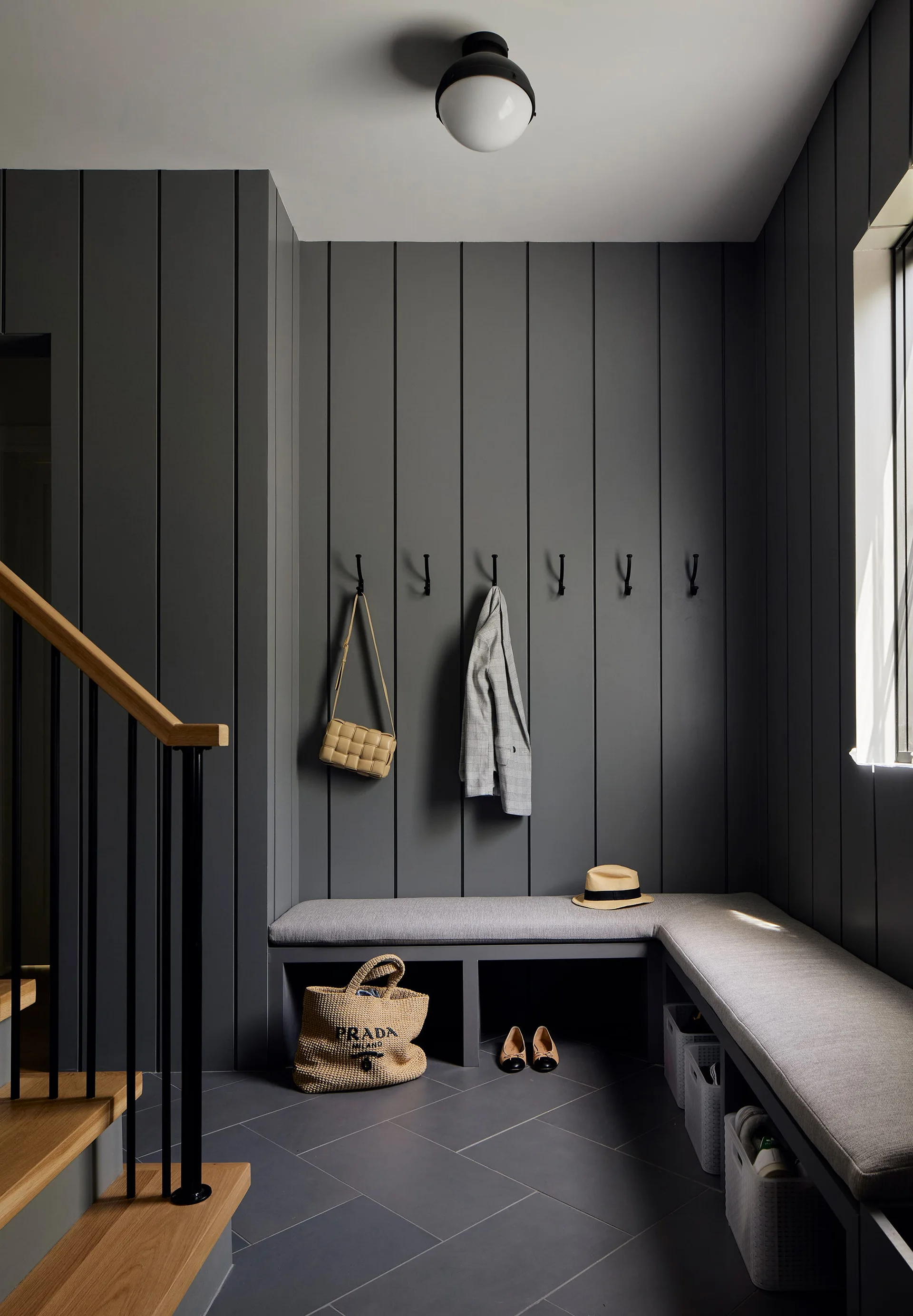A dark charcoal mudroom features vertical wood paneling, black wall hooks, a built-in L-shaped bench with cushions, and tiled flooring.