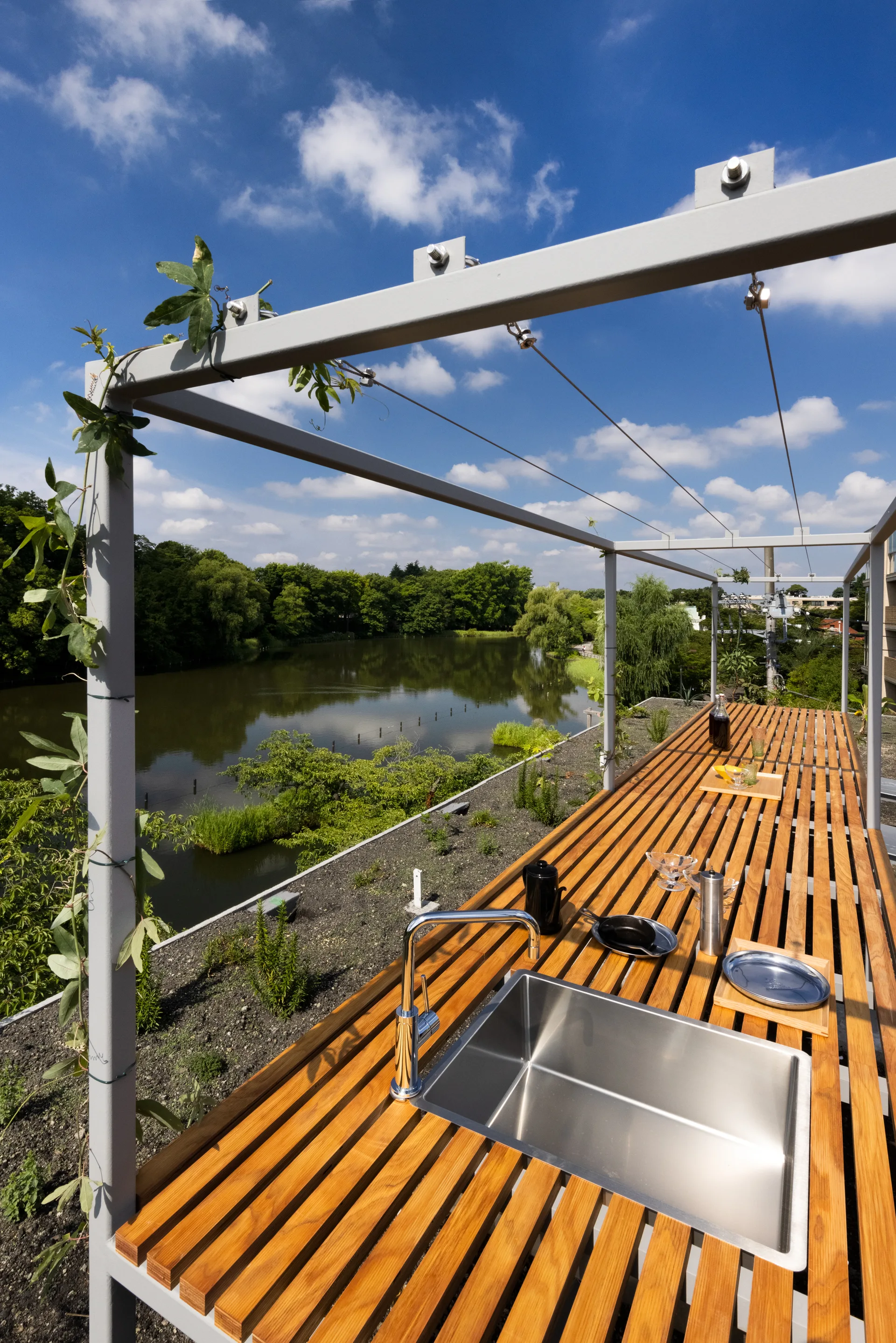 A modern outdoor kitchen features a long slatted wooden countertop and a stainless steel sink under a metal pergola overlooking a lake.