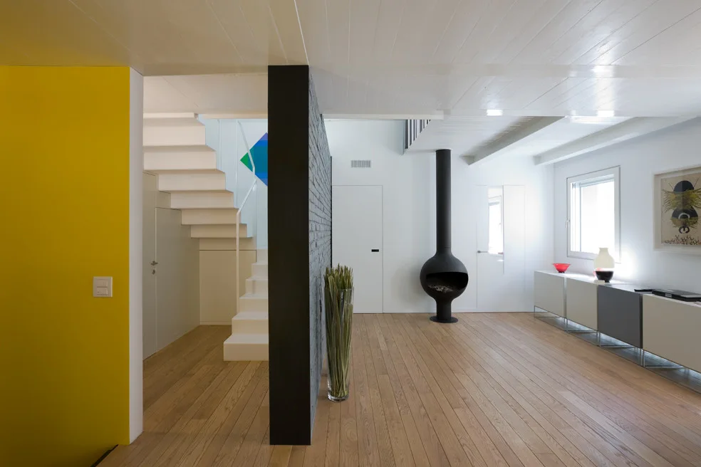 A minimalist living area with light wood flooring features a dark grey brick partition wall, a white staircase, and a black spherical fireplace.