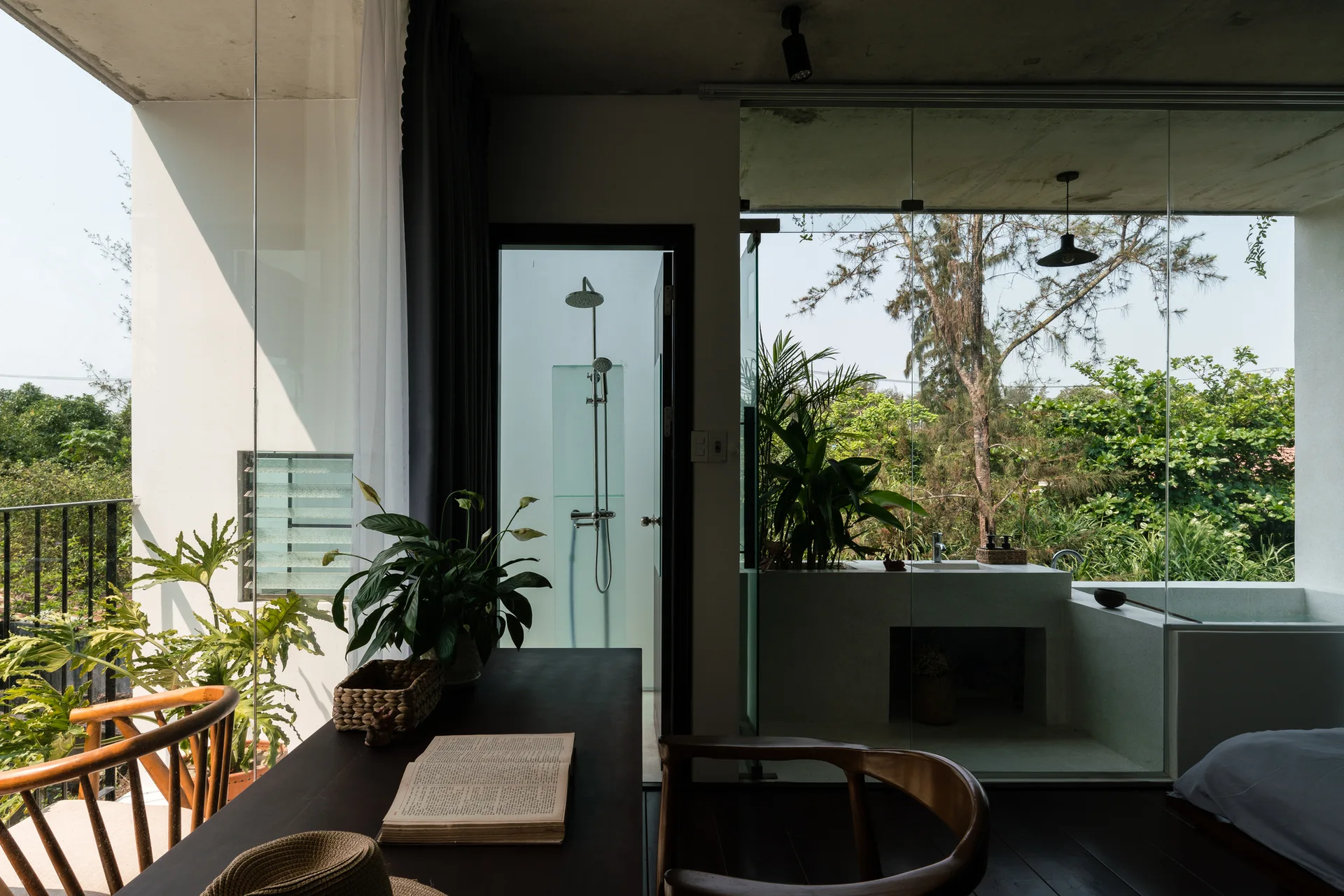 A modern bedroom featuring a dark wood desk, raw concrete ceiling, and large glass walls looking out to an open bathroom and lush garden.