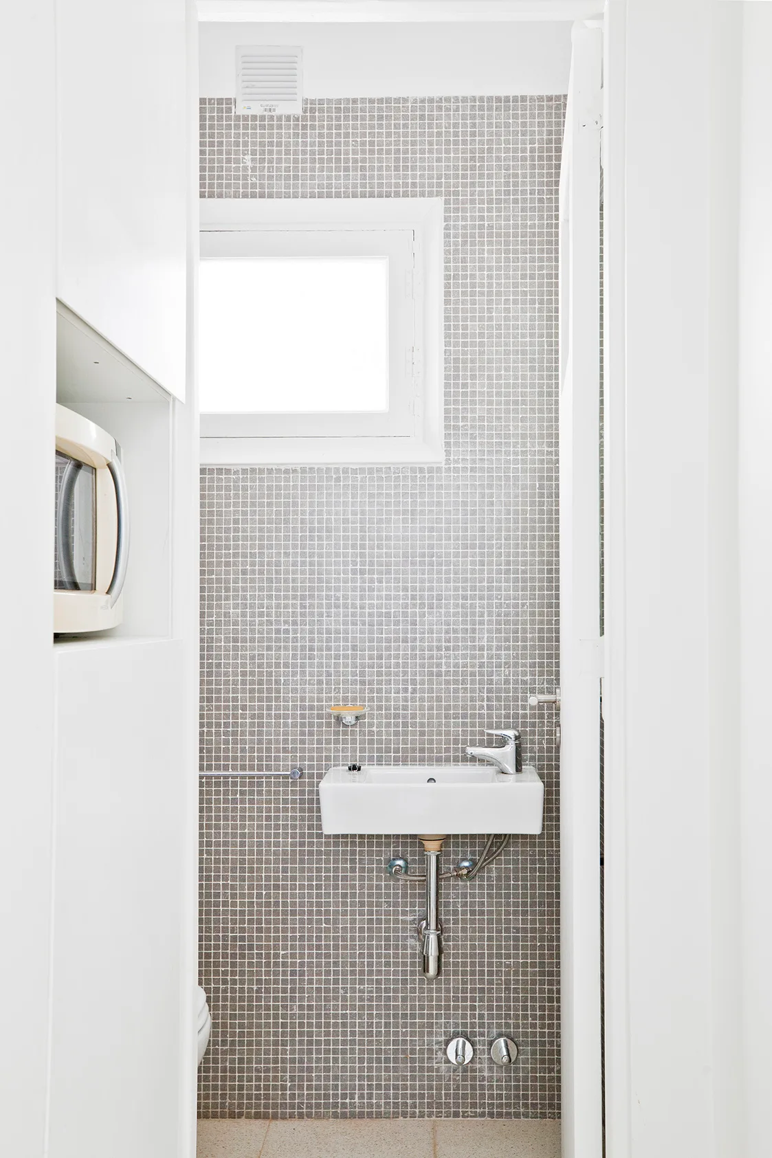 A narrow white bathroom features a grey mosaic tile wall, a small wall-mounted sink, and a high window.