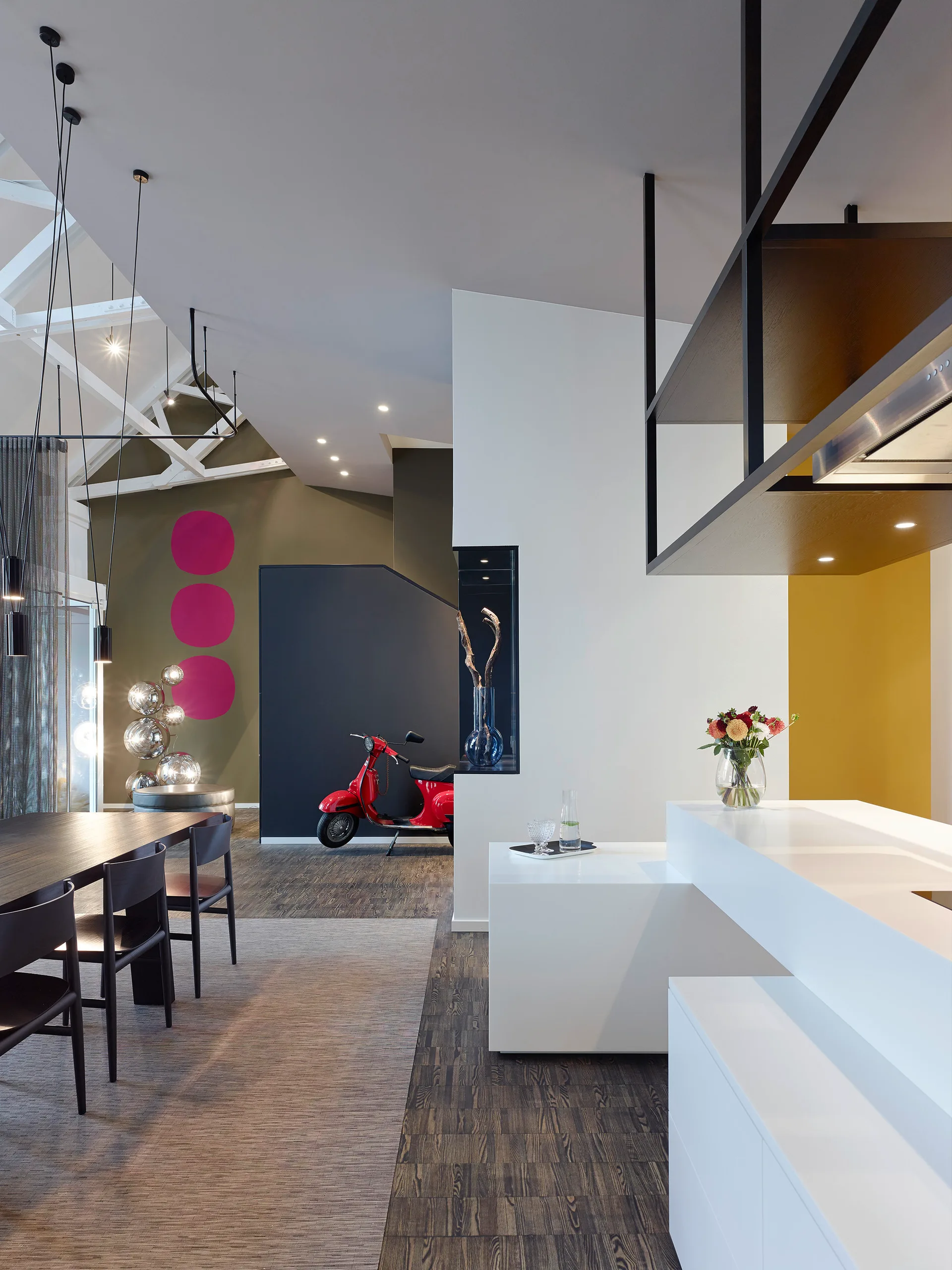 A modern open-concept kitchen and dining area featuring a tiered white island, dark wood table, exposed white ceiling trusses, and a red scooter.