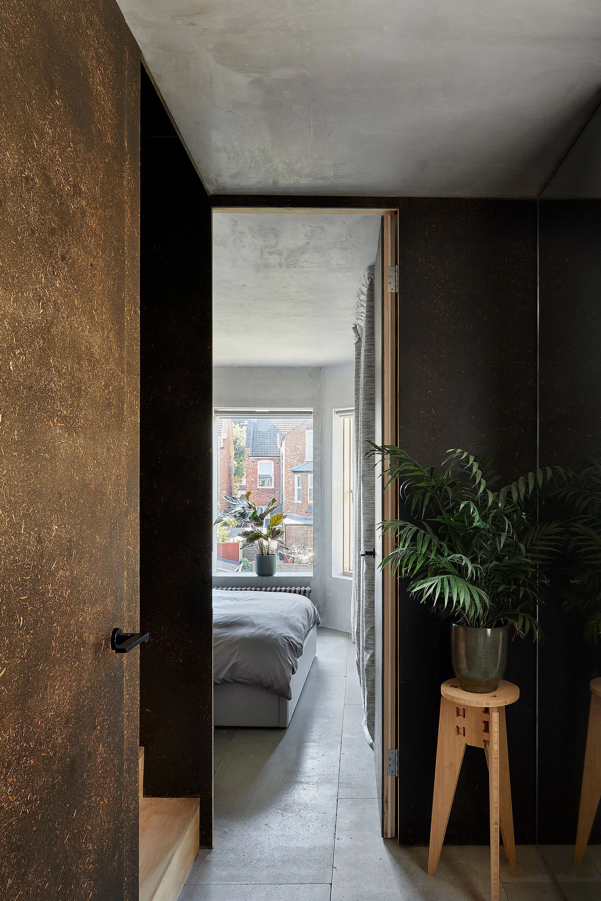 A view through a dark-paneled doorway into a minimalist bedroom with a grey bed, concrete ceiling, and a potted plant on a wooden stool.