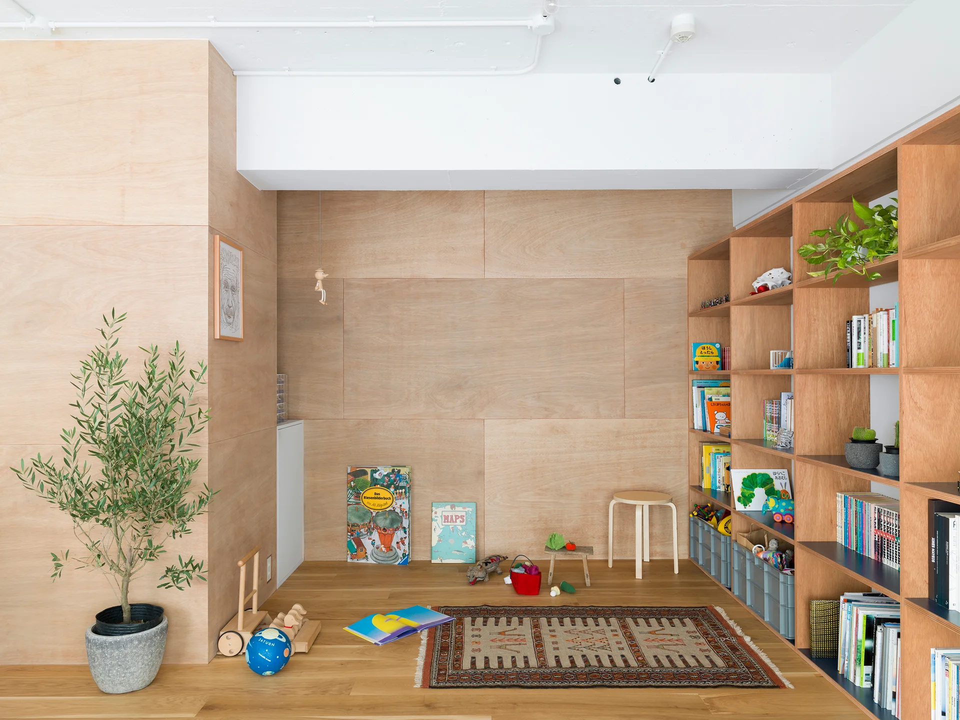A kids playroom with light plywood walls, a matching wooden bookshelf filled with books, a patterned area rug, and wood flooring.