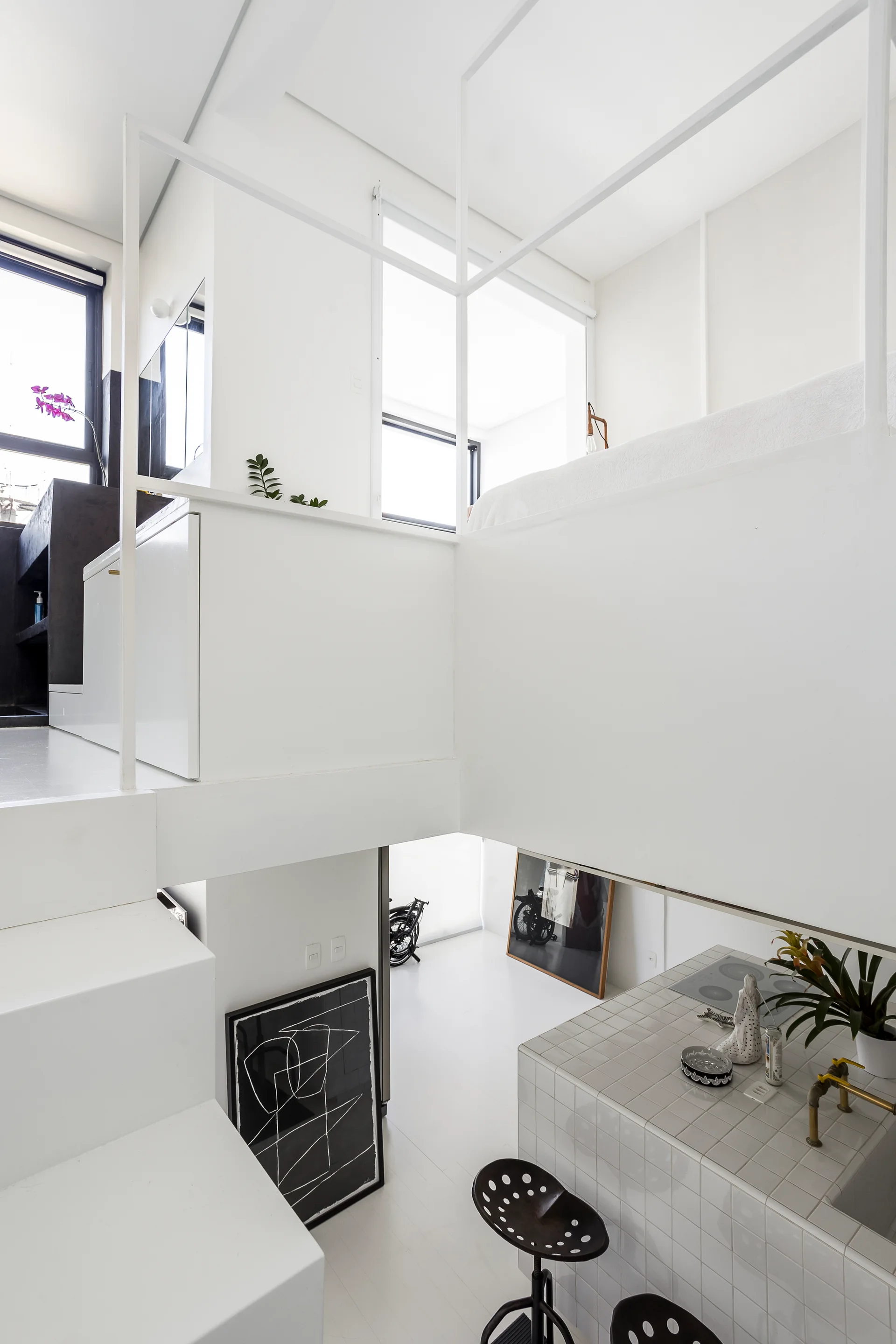 A split-level white minimalist loft featuring a tiled kitchen island, black metal stools, a white staircase, and an upper-level sleeping area.