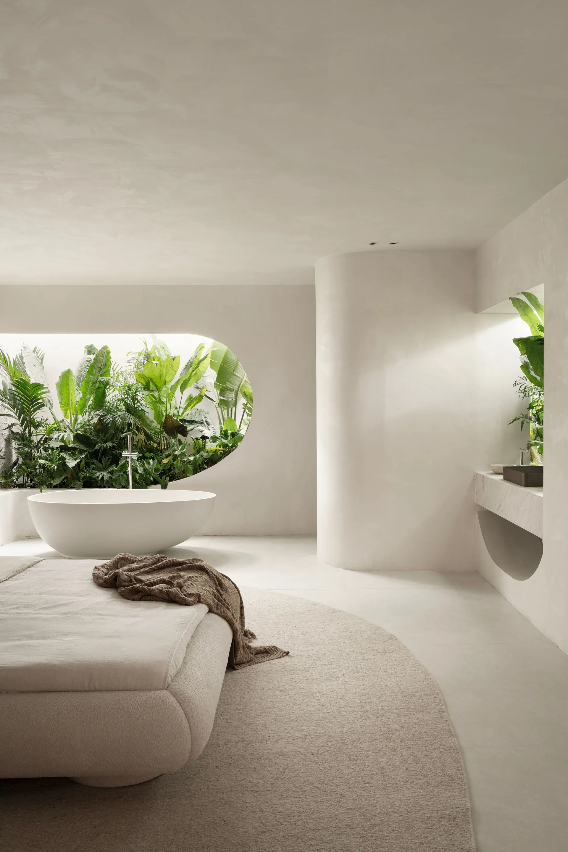 A minimalist off-white bedroom featuring a low bed, a curved plaster wall, an integrated vanity, and an oval window showing a bathtub nestled in plants.