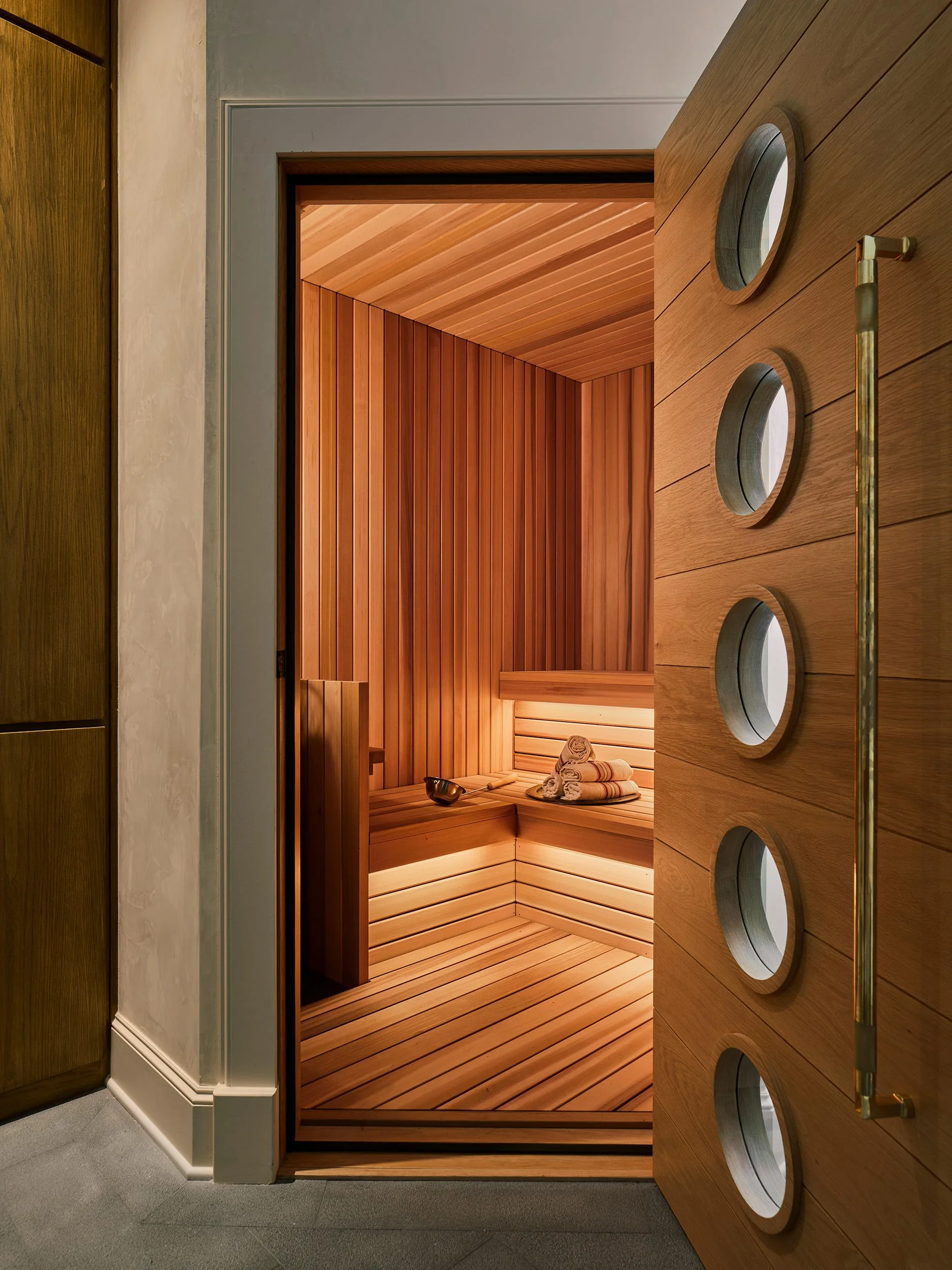 An open wooden sauna door with five circular windows reveals a cedar-lined interior with tiered benches and soft under-bench lighting.