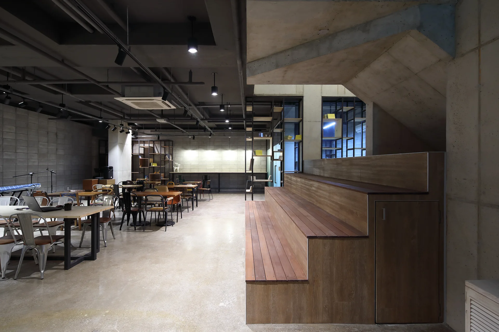 An open industrial room featuring concrete walls, a tiered wooden seating platform, and scattered cafe tables and chairs under an exposed ceiling.