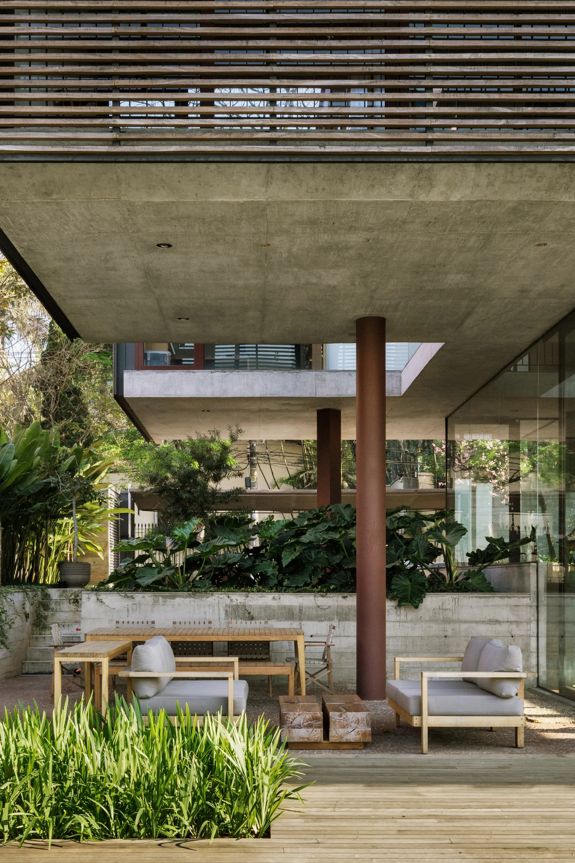 A modern outdoor patio features gray upholstered lounge chairs and a wooden dining set beneath a large concrete overhang and lush green plants.