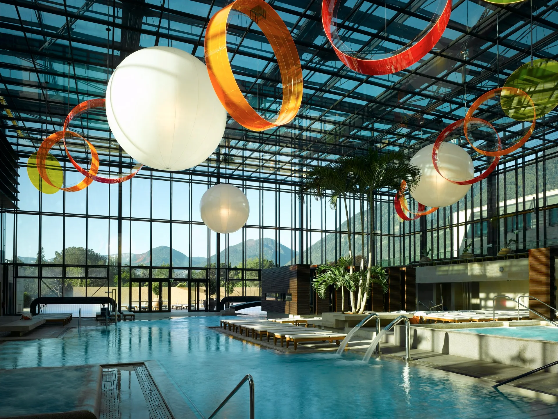 An indoor swimming pool inside a glass atrium features oversized white sphere pendant lights, colorful hanging ring sculptures, and rows of sun loungers.