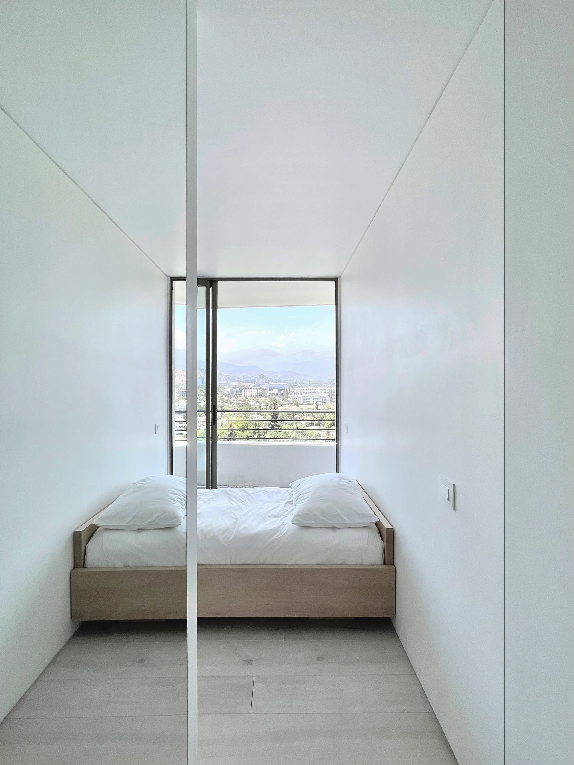 A minimalist white bedroom features a wooden platform bed fit tightly between walls, looking out a large window toward a city view.