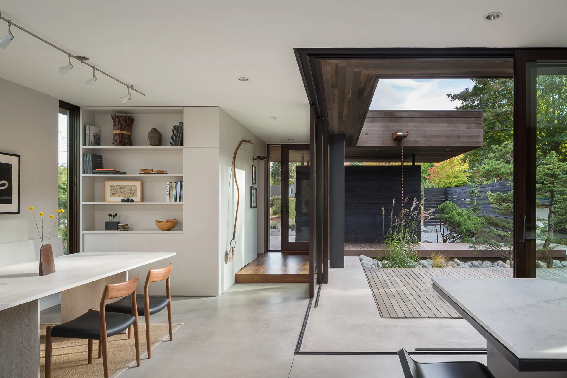 A modern dining room features a light wood table, minimalist chairs, and built-in shelving next to sliding glass doors that open to a wood deck.