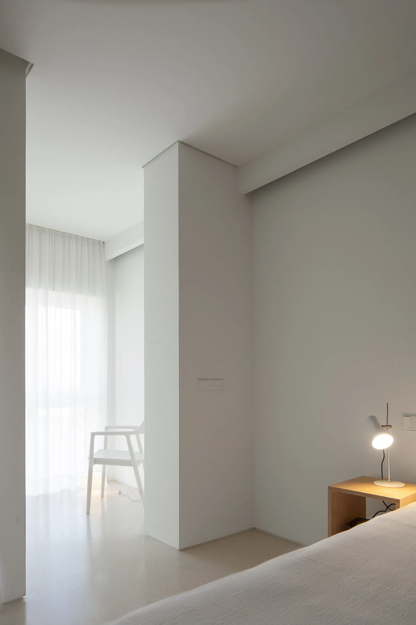 A minimalist white bedroom features a light wood bedside table with a glowing lamp, and a white chair near a window with sheer curtains in the background.