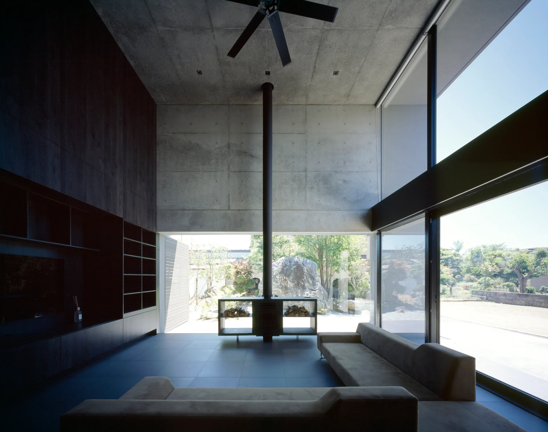 A modern living room features low-profile grey sofas, a central black freestanding fireplace, exposed concrete walls, and large floor-to-ceiling windows.