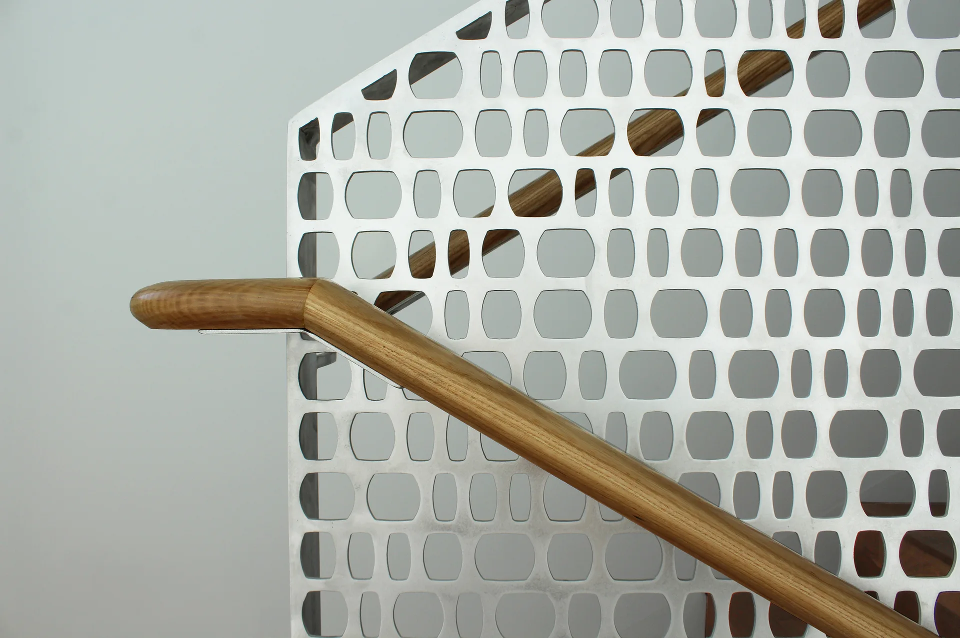 A close-up of a modern staircase railing featuring a smooth wooden handrail mounted on a silver perforated metal screen with a geometric grid pattern.