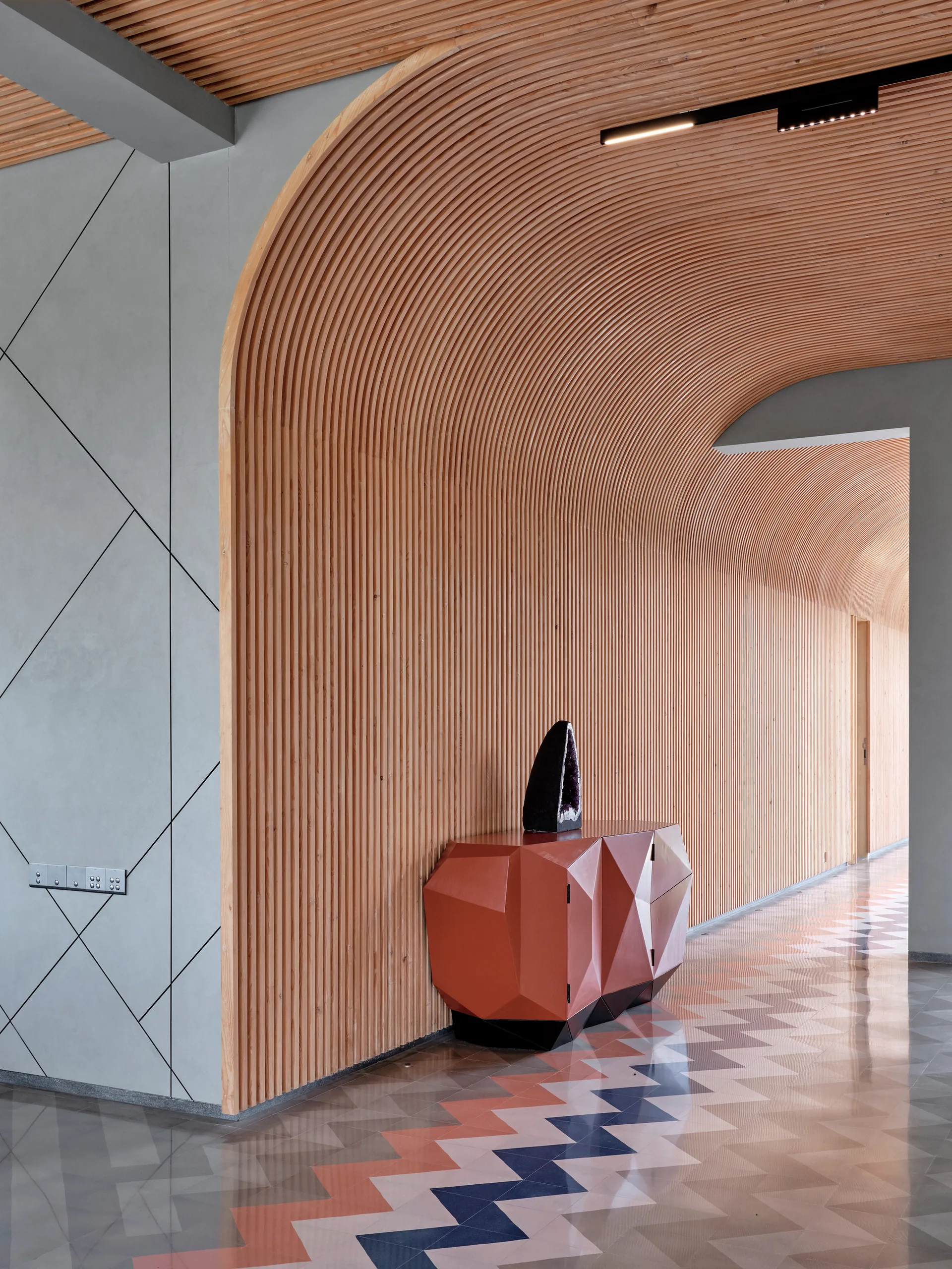 A modern hallway features a curved slatted wood vaulted ceiling, a faceted orange credenza, and grey geometric wall paneling.