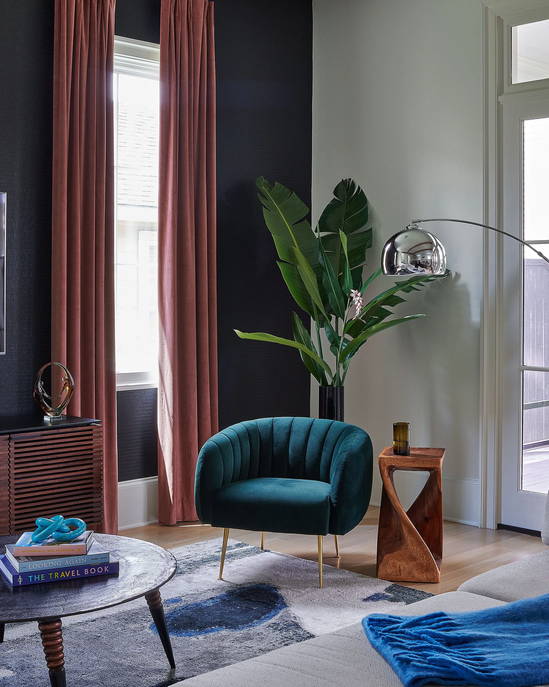 A channel-tufted teal velvet armchair sits on an abstract rug next to a twisted wood side table, framed by terracotta curtains and a dark wall.