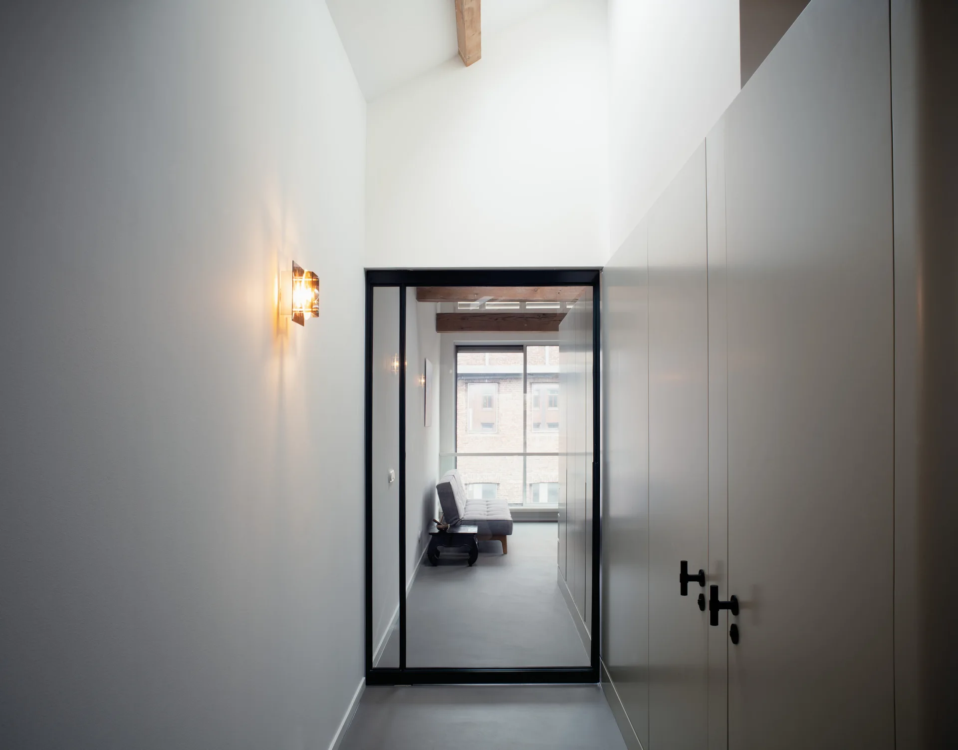 A minimalist white hallway features built-in cabinets, a warm wall sconce, and a black-framed glass door looking into a room with wooden beams.