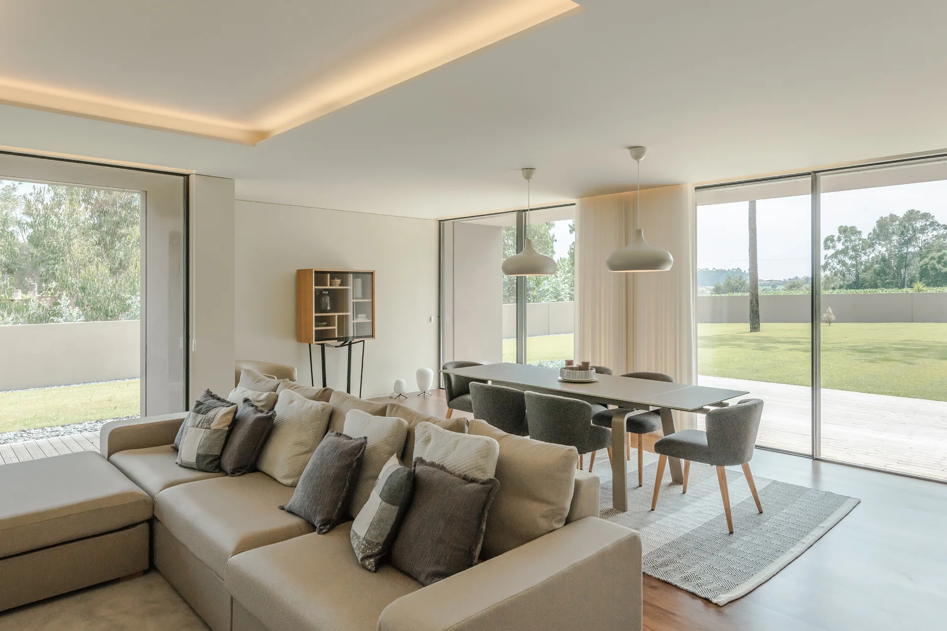 A modern open-plan living and dining space features a large beige sectional sofa, a grey dining set, and floor-to-ceiling glass doors.