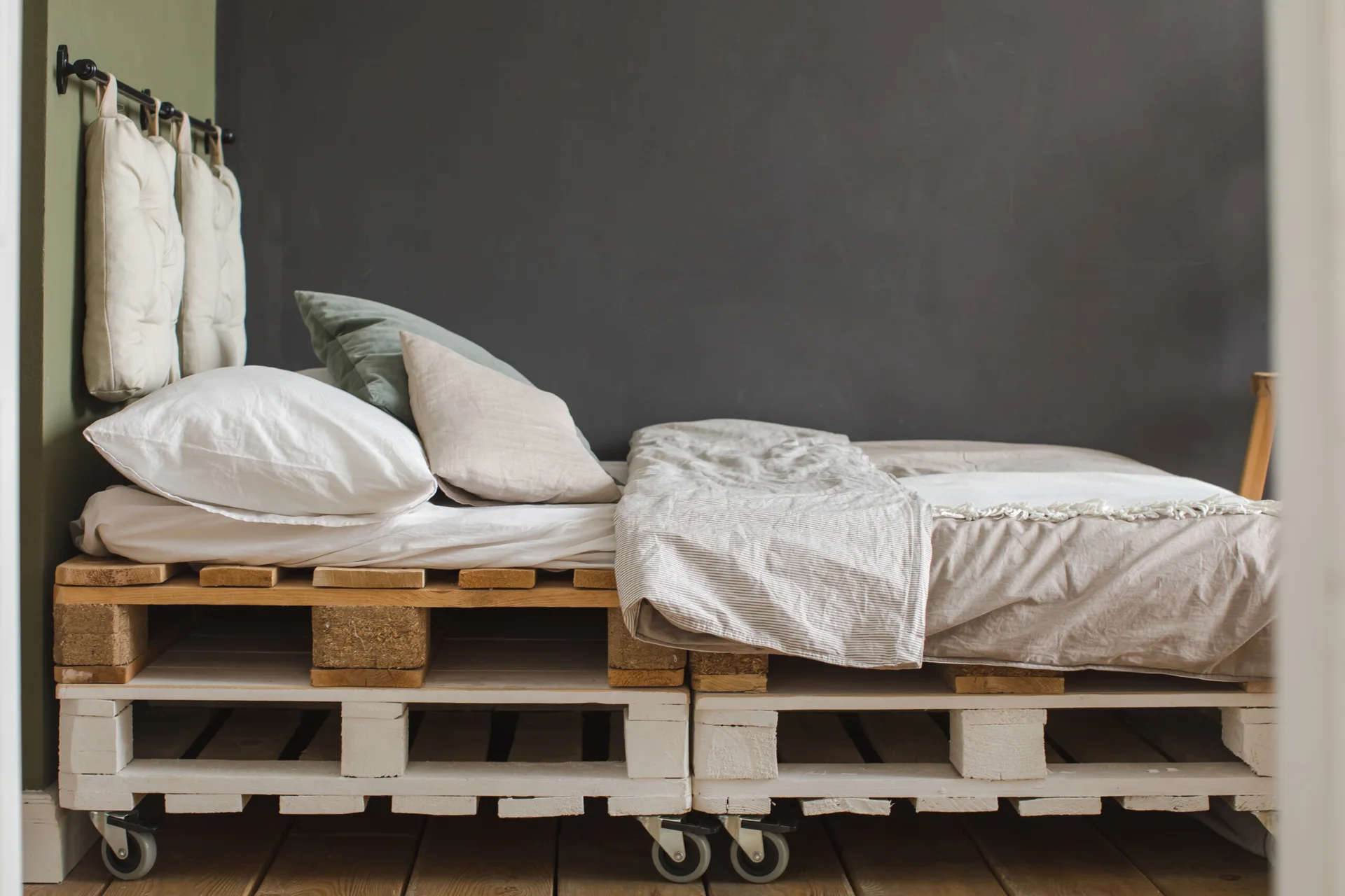 A low-profile bed made from stacked wooden pallets on wheels features hanging white cushions as a headboard against a dark grey wall.