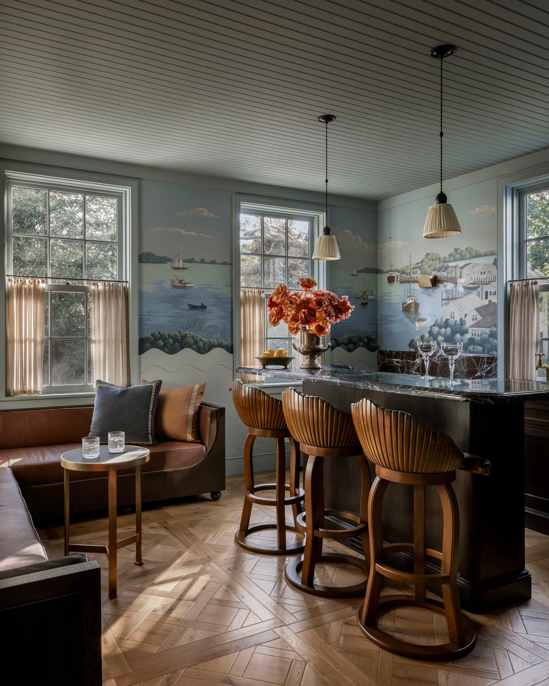 A home bar featuring a scenic coastal mural, three wooden shell-back stools, a dark stone counter, and a brown leather sofa on parquet flooring.