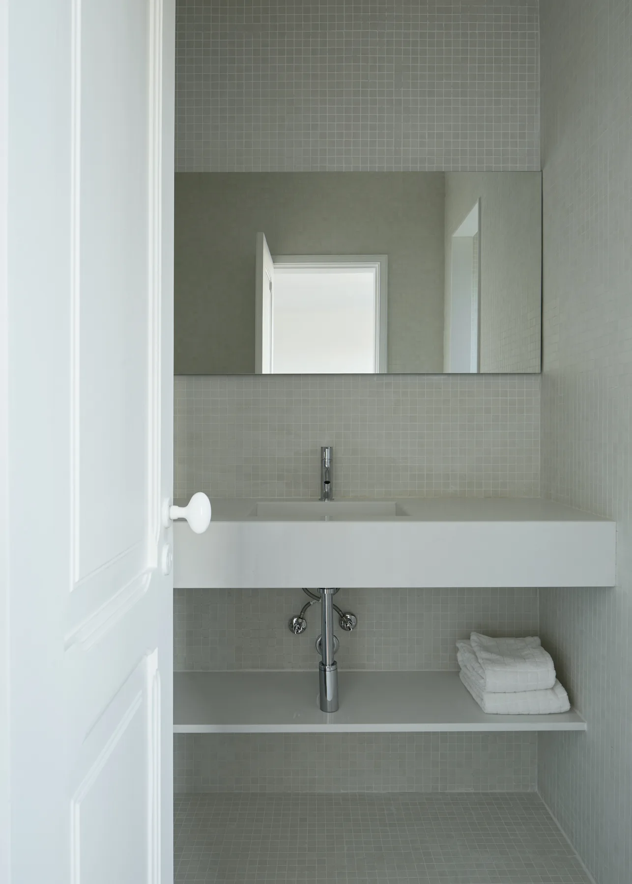 A white floating vanity with a chrome faucet stands against light grey mosaic tile walls and floors in a minimalist bathroom.