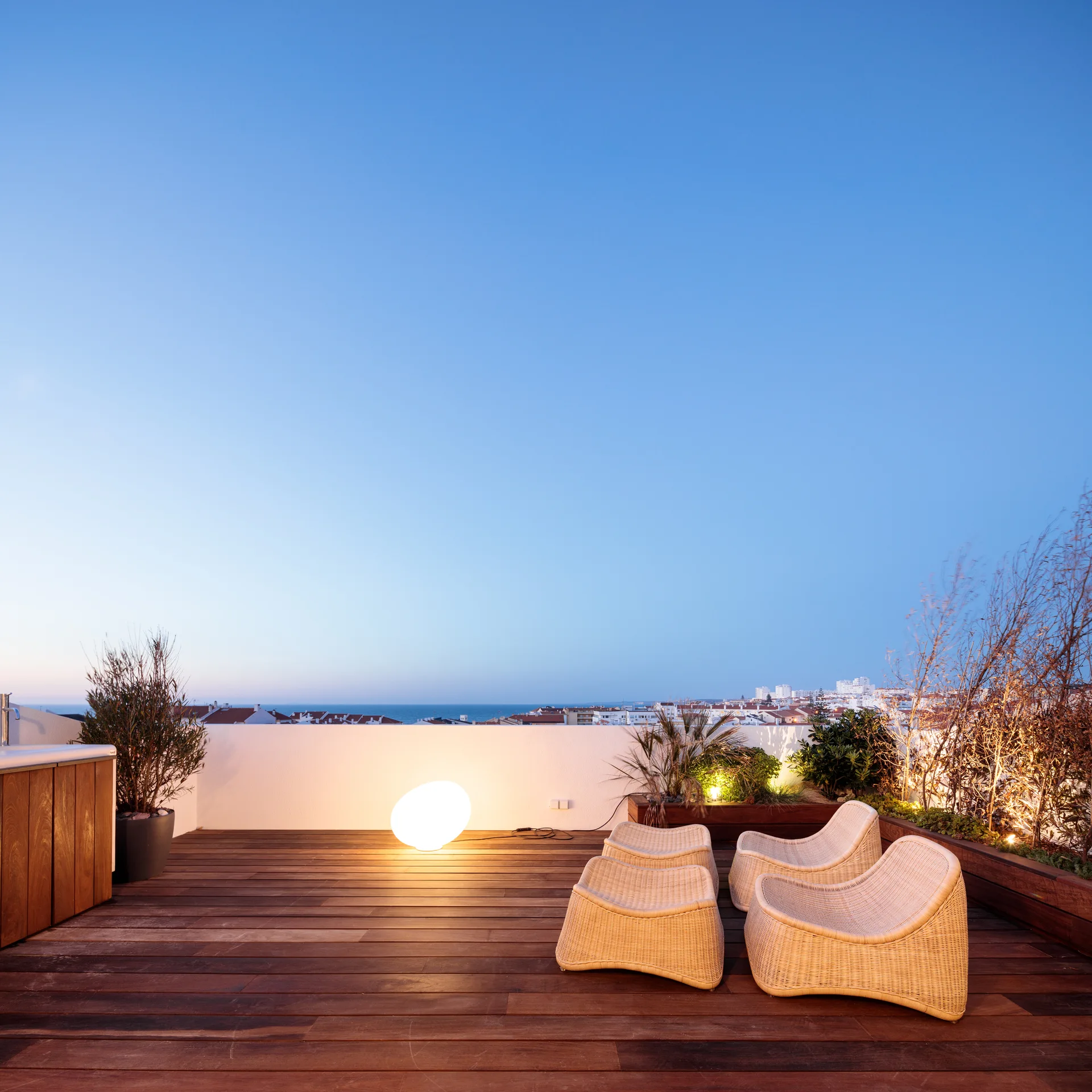 Two woven wicker lounge chairs and ottomans sit on a wooden rooftop deck beside a glowing orb lamp overlooking a distant city skyline.