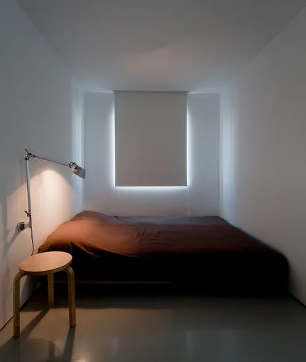 A minimalist narrow bedroom with a low-profile bed, white walls, a wooden stool nightstand, and an adjustable wall-mounted lamp.