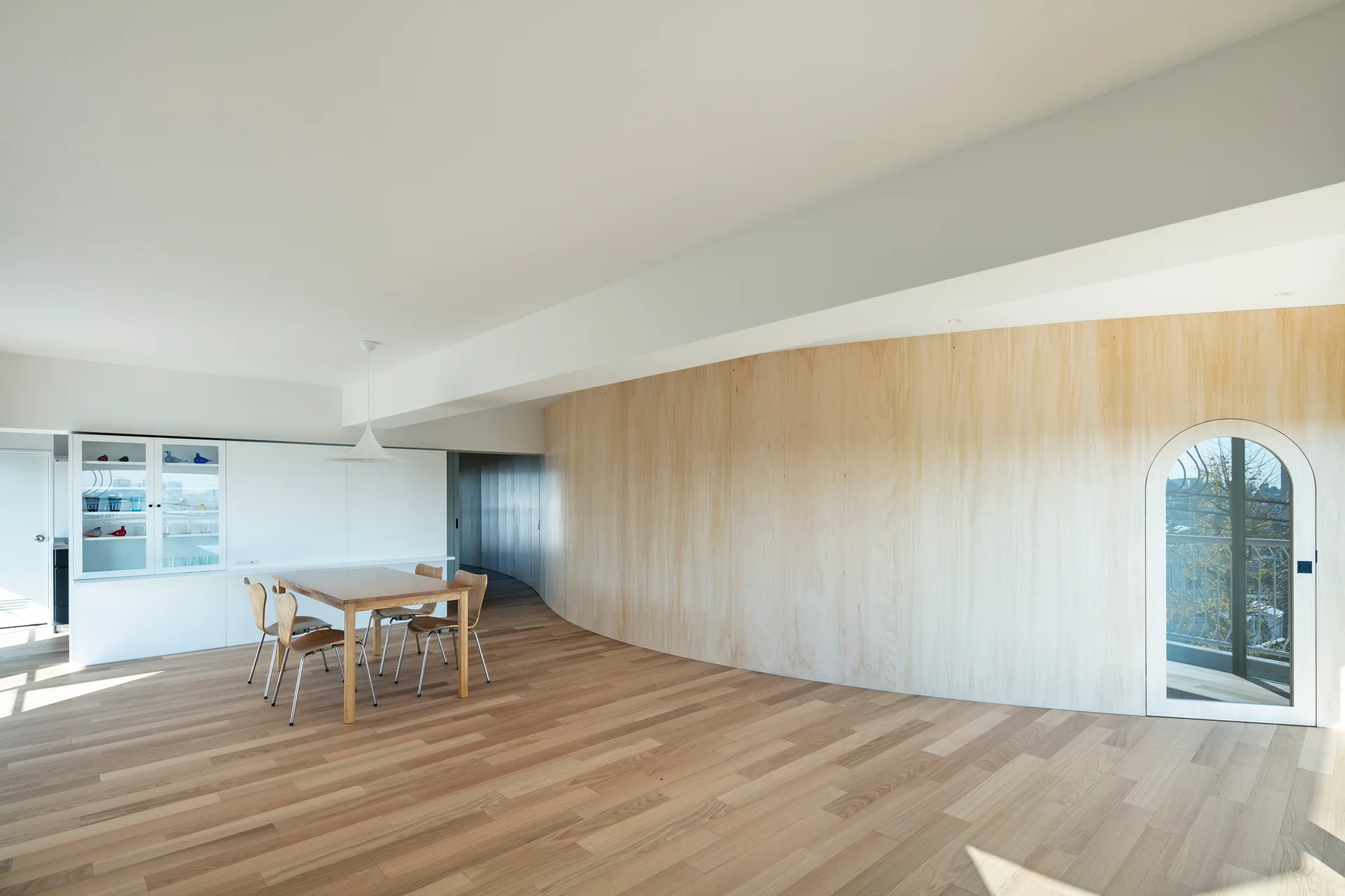 A minimalist dining room features a wooden table and chairs, a large curved plywood wall, and an arched glass door leading outside.