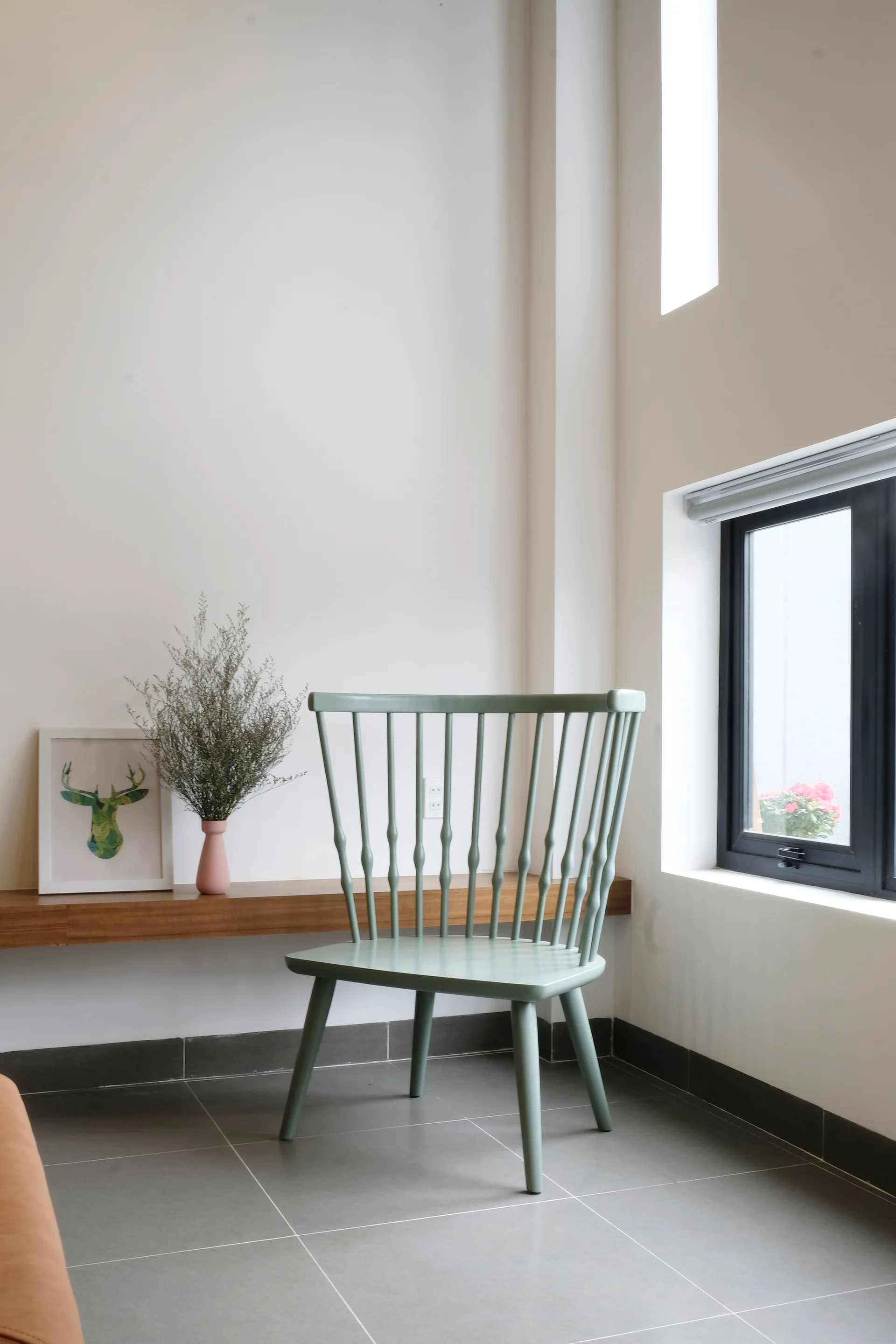 A sage green spindle-back chair sits on gray tile flooring next to a floating wooden shelf with a pink vase and framed art.