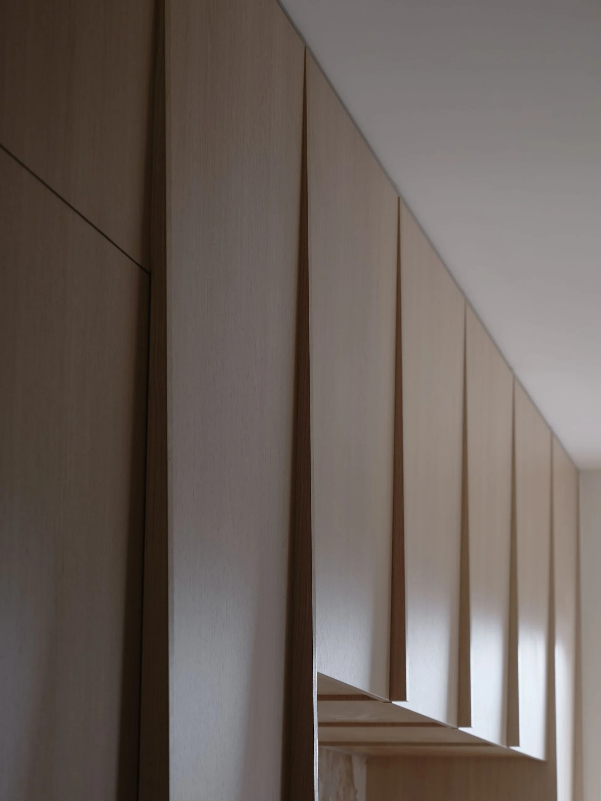 A close-up view of minimalist light wood kitchen cabinet doors with integrated tapered vertical handles beneath a plain white ceiling.