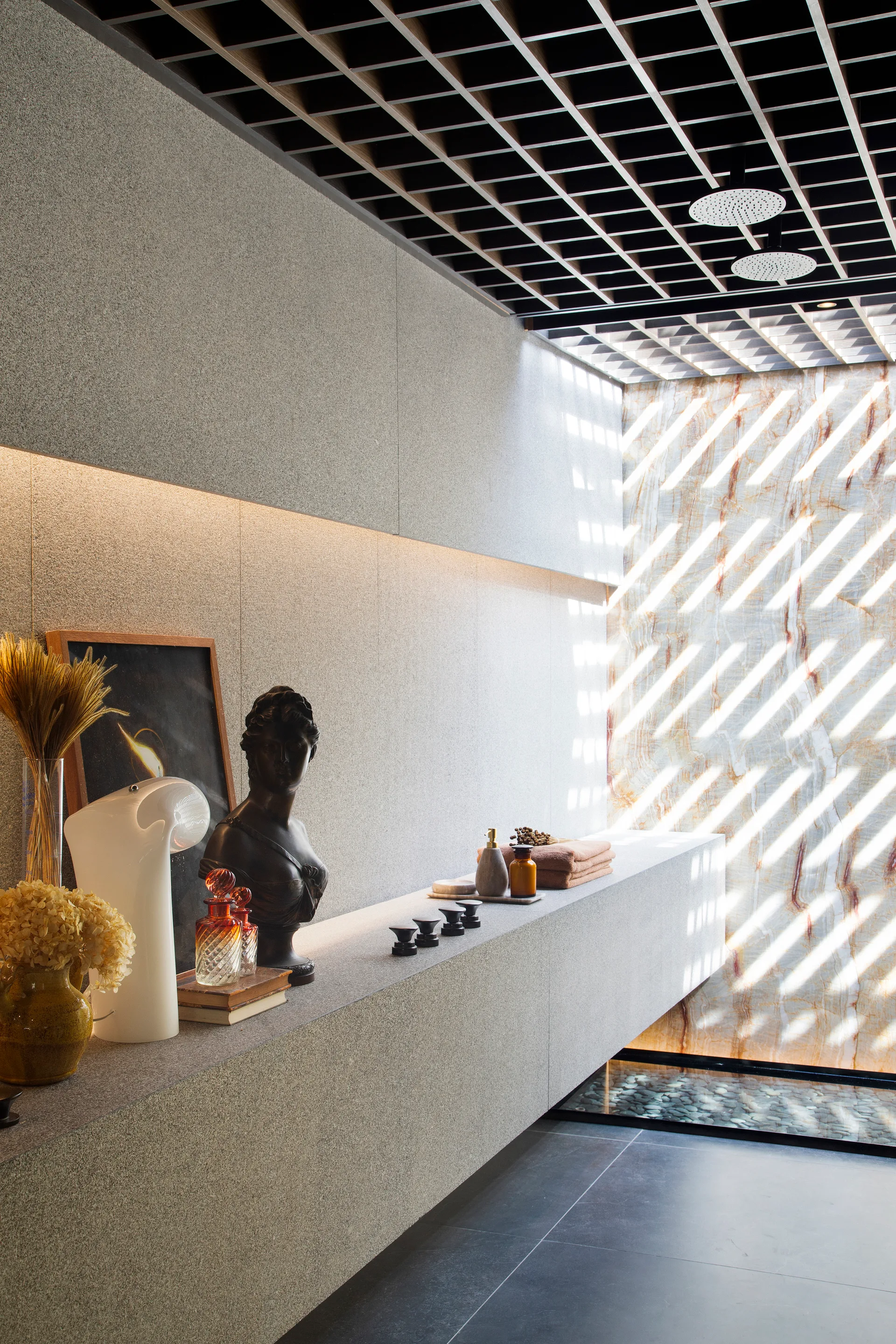 A grey stone bathroom features a long floating vanity, a black grid ceiling with dual shower heads, and patterned light cast on a marble wall.