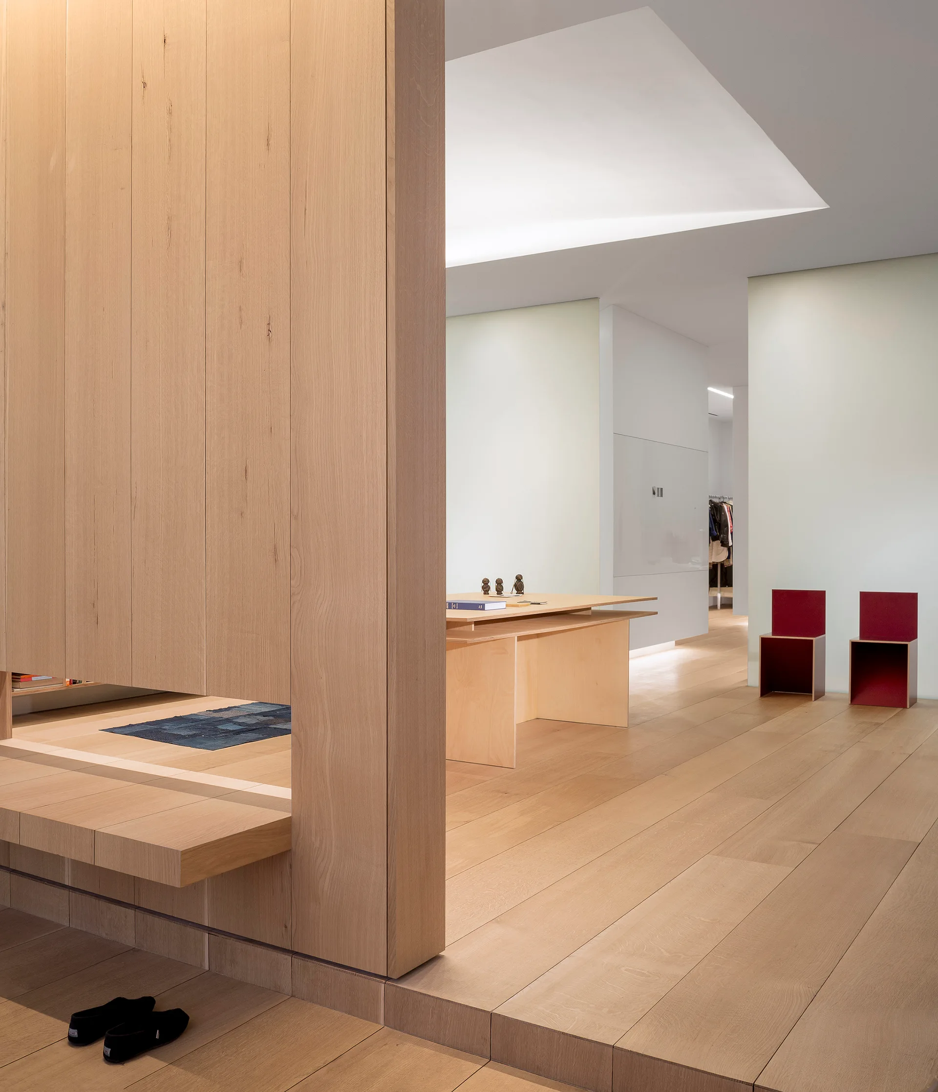 A minimalist home office features light wood plank flooring, a large oak partition wall, a matching wooden desk, and two red cube chairs.