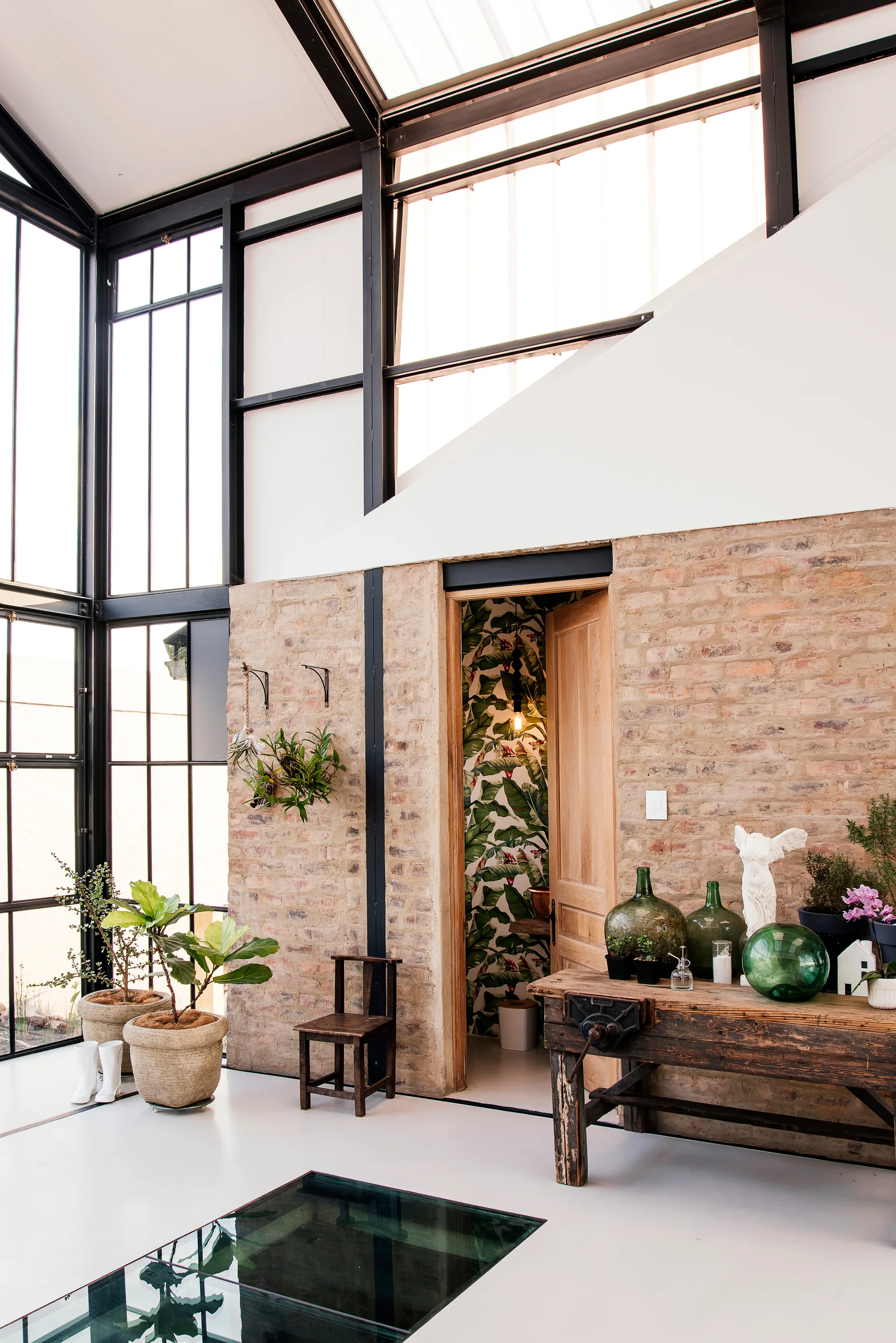 A sunlit conservatory features exposed brick walls, a weathered wooden workbench, and a doorway revealing green botanical wallpaper.