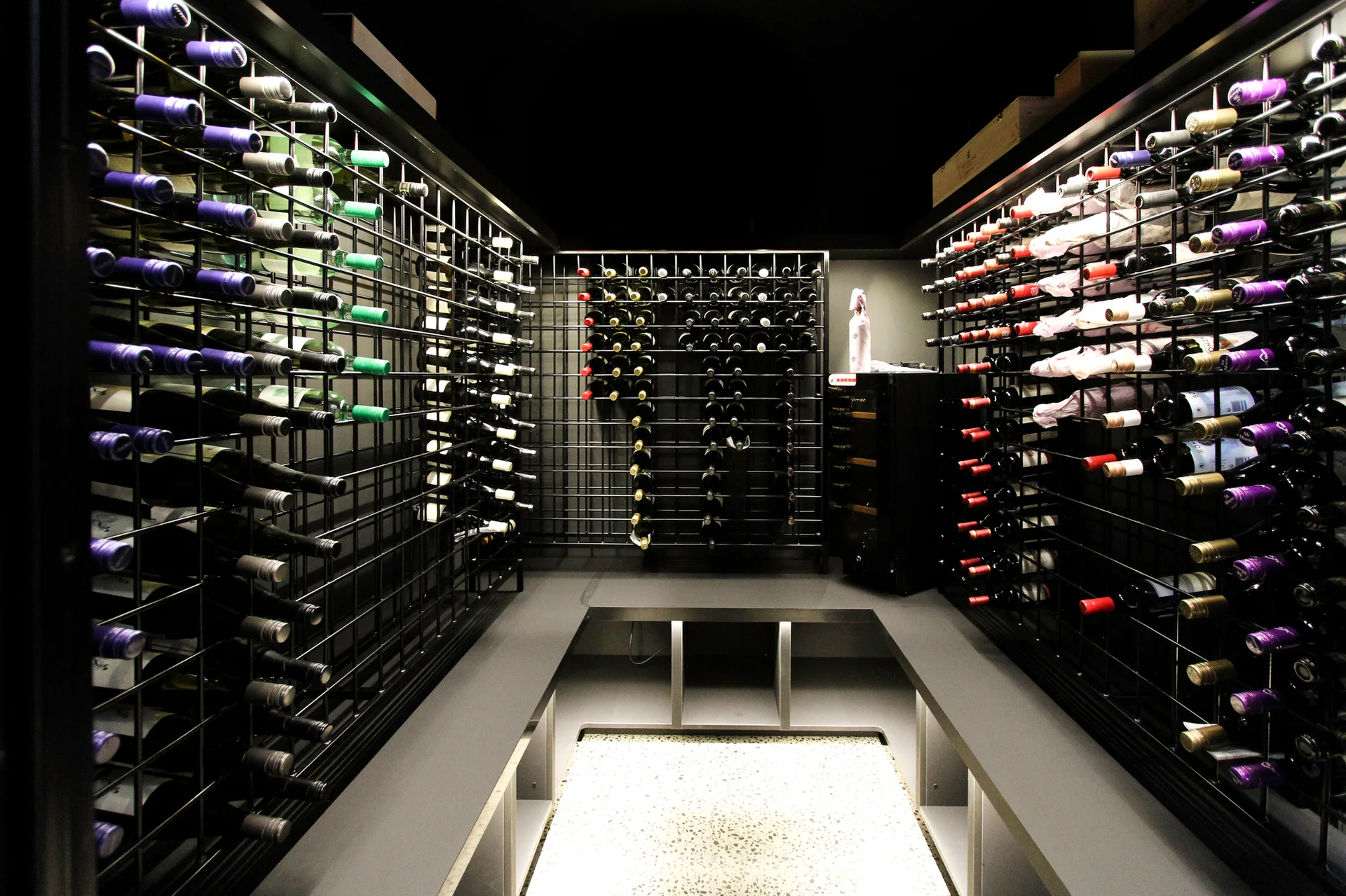 A symmetrical wine room featuring black metal grid racking filled with wine bottles, dark grey countertops, and a light speckled floor.