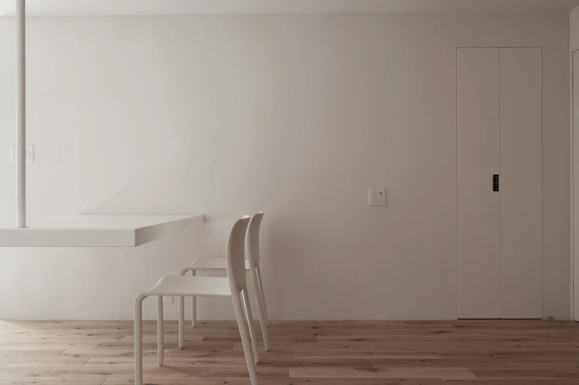 A minimalist dining area featuring a white floating table, matching white chairs, and light wood flooring against a plain wall.