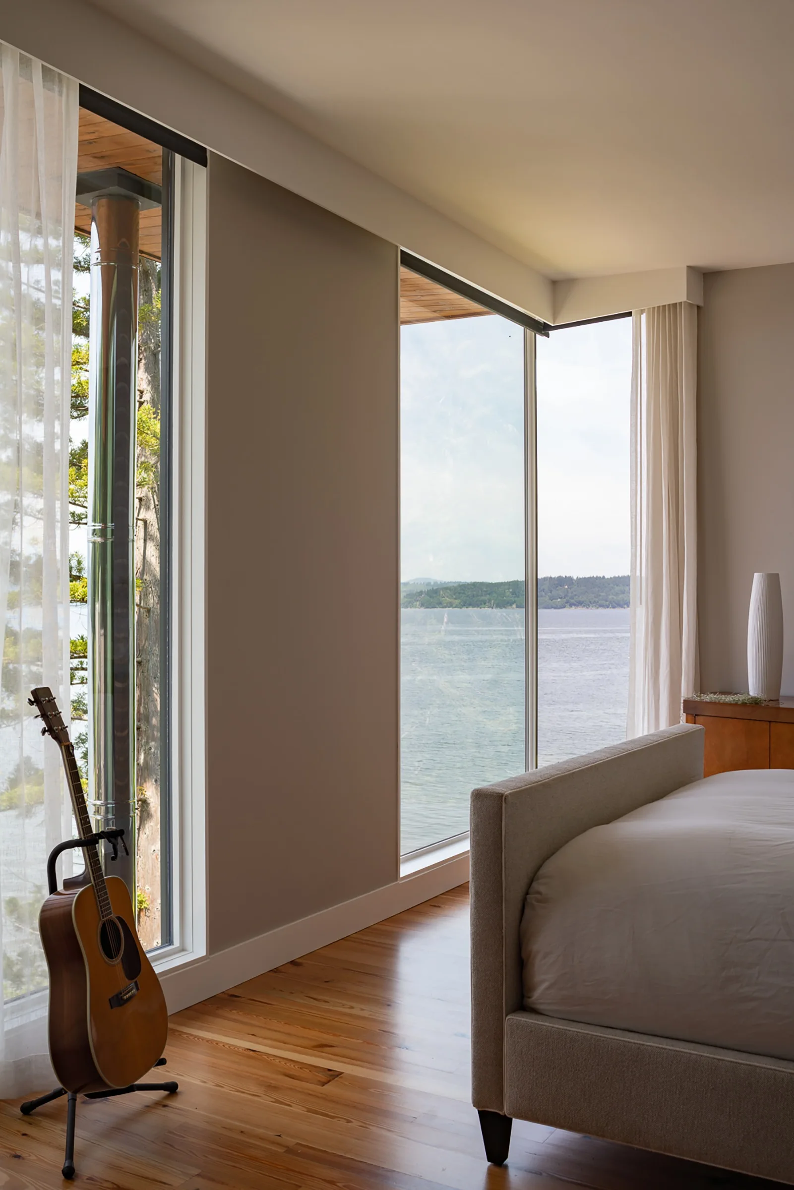 A minimalist bedroom featuring an upholstered bed, hardwood floors, and large windows that overlook a scenic water view.