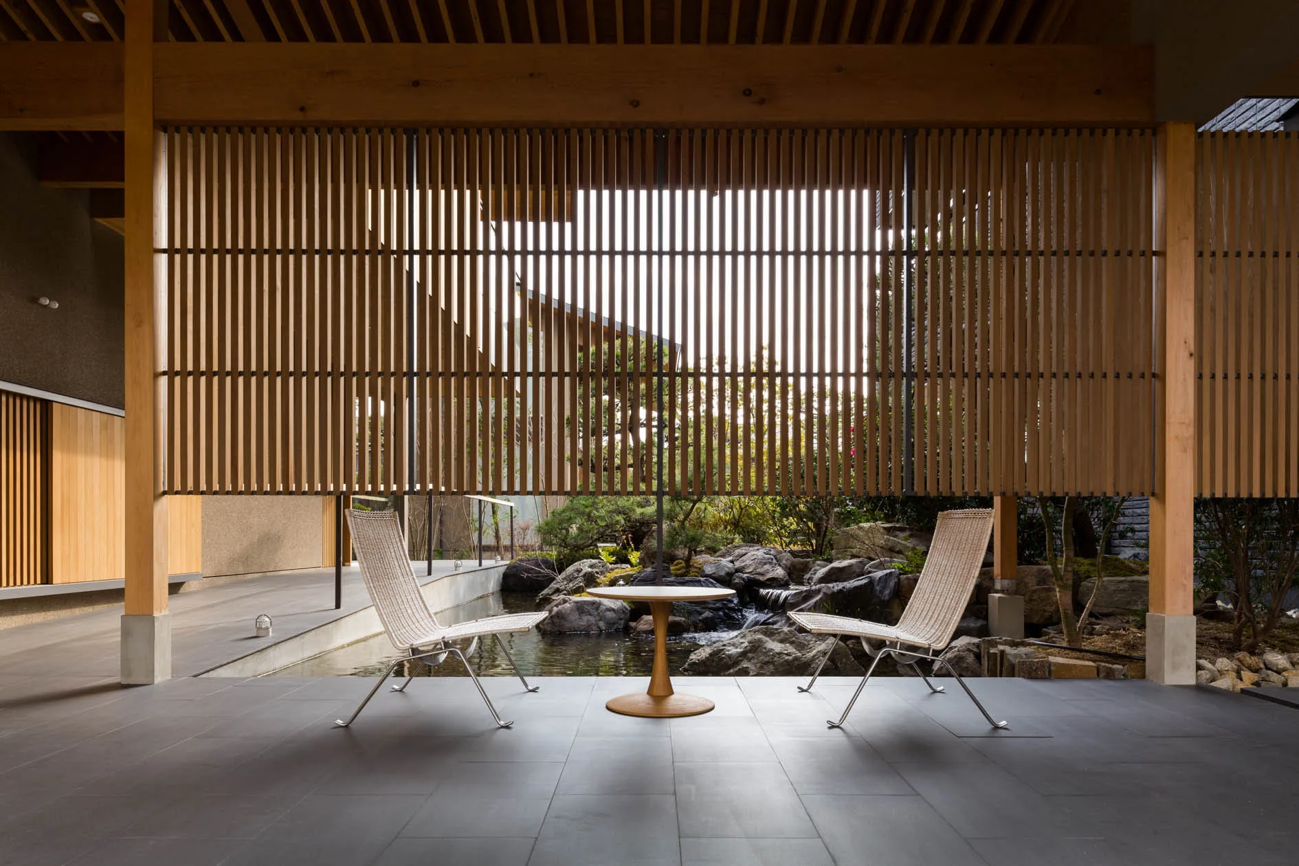 Two lounge chairs and a small table sit on a dark tiled patio beneath a slatted wood screen overlooking a rock water garden.