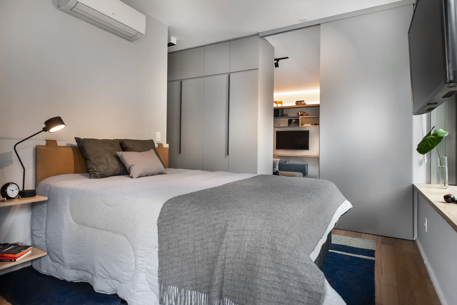 A minimalist gray bedroom features a bed with a tan leather headboard, a blue striped rug, and built-in wardrobes with a sliding partition door.