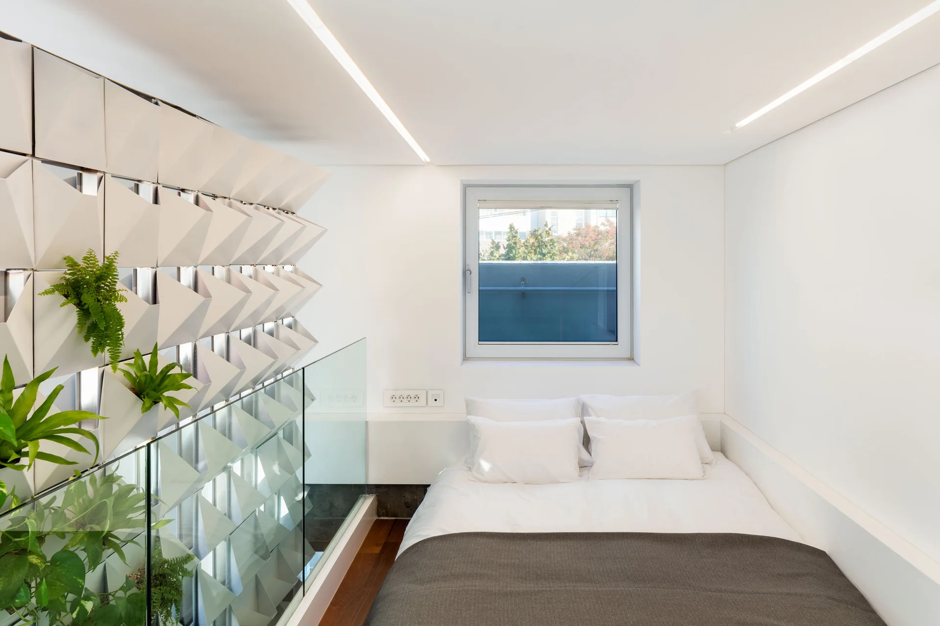A minimalist white bedroom with a bed, a glass partition railing, and a textured geometric wall installation holding leafy green plants.