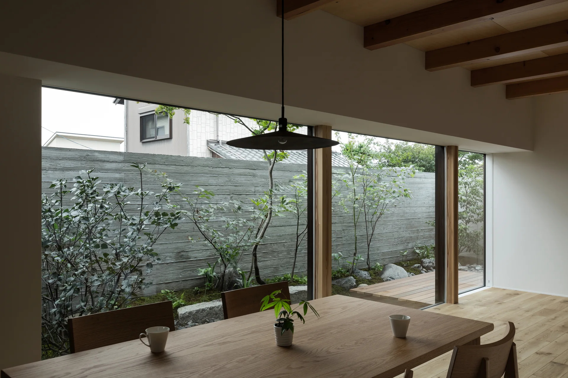 A minimalist dining room features a wooden table, exposed timber ceiling beams, and a large window facing a small garden and concrete wall.