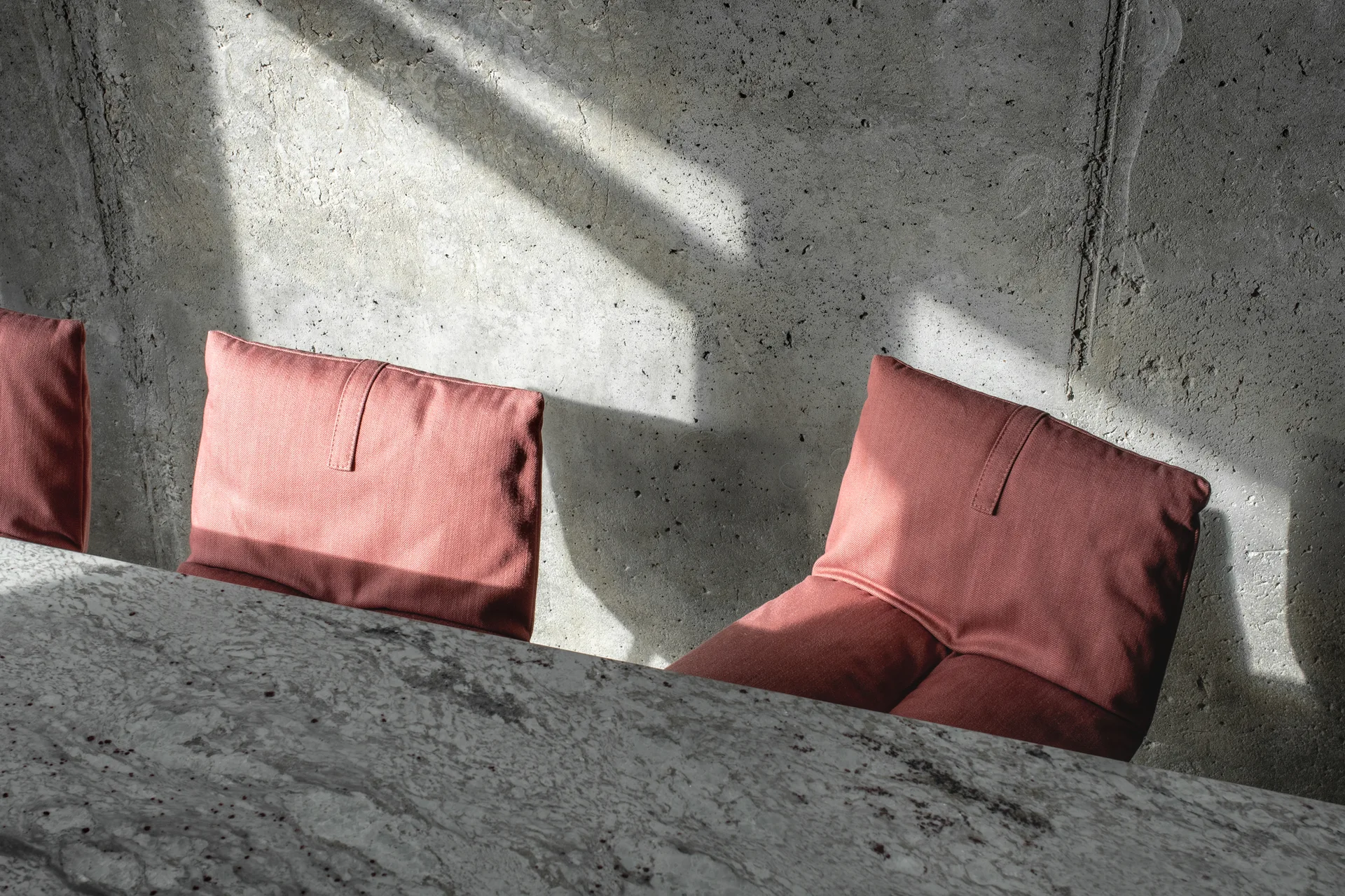 Rose-colored upholstered chairs stand beside a marble table against a textured grey concrete wall illuminated by sharp sunlight.